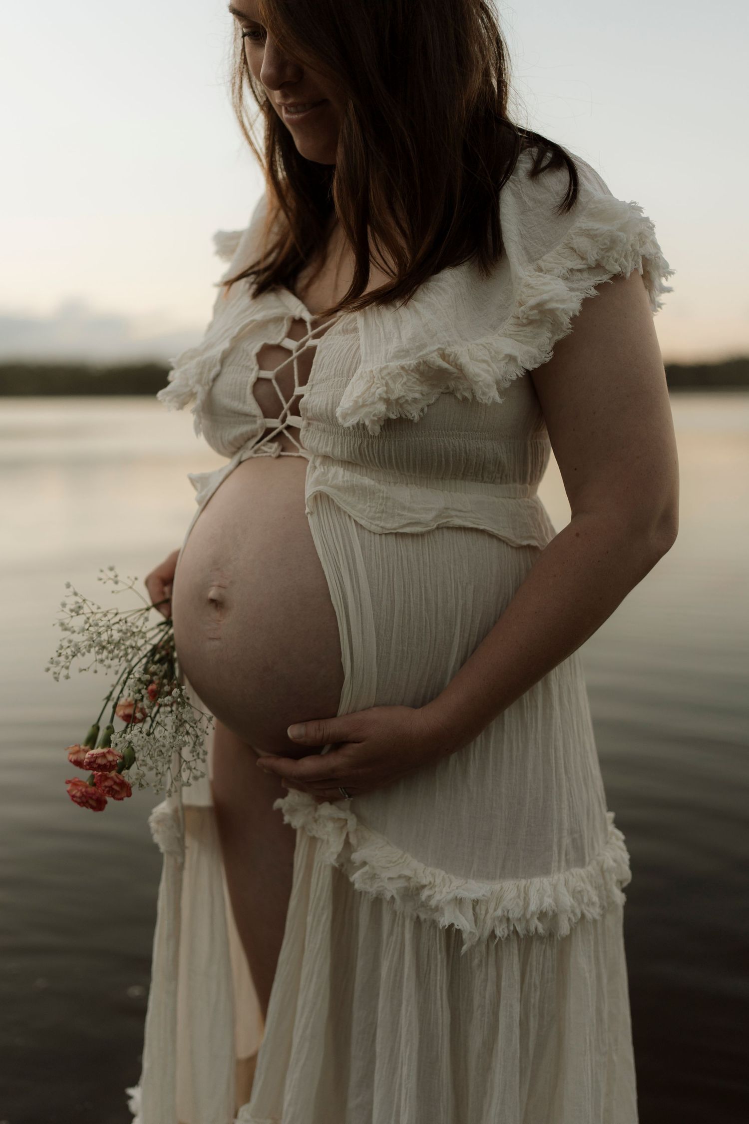 Pregnant woman in white flowy dress holding delicate flowers on beach at sunset, soft ethereal maternity photography.
