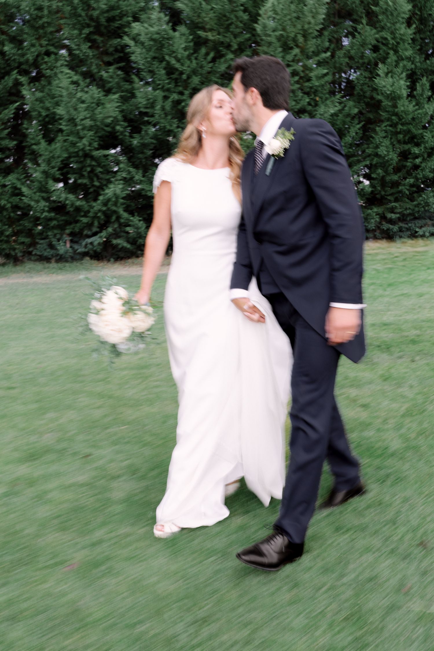 A couple in wedding attire share a romantic moment while walking on green grass with evergreen trees in the background.