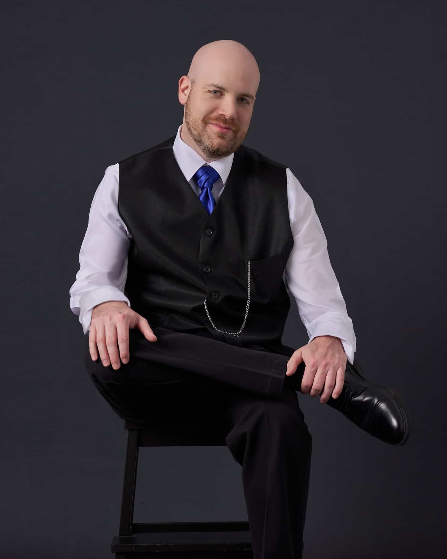 Thirty-something bald man in dark suit and tie seated with leg crossed on stool in photo session in Perry Hall Maryland