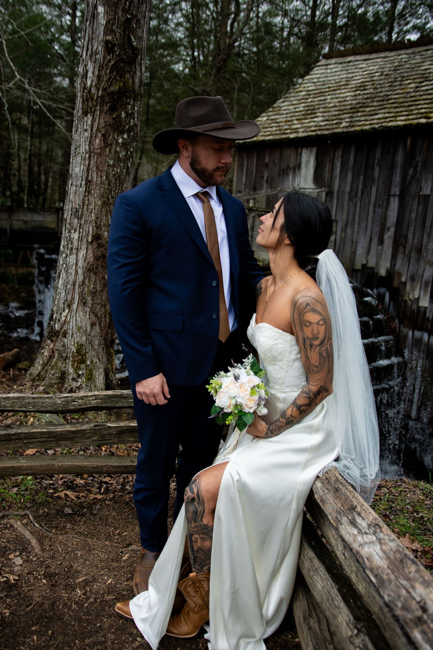 A couple in wedding attire shares an intimate moment in a rustic outdoor setting among trees and wooden structures.