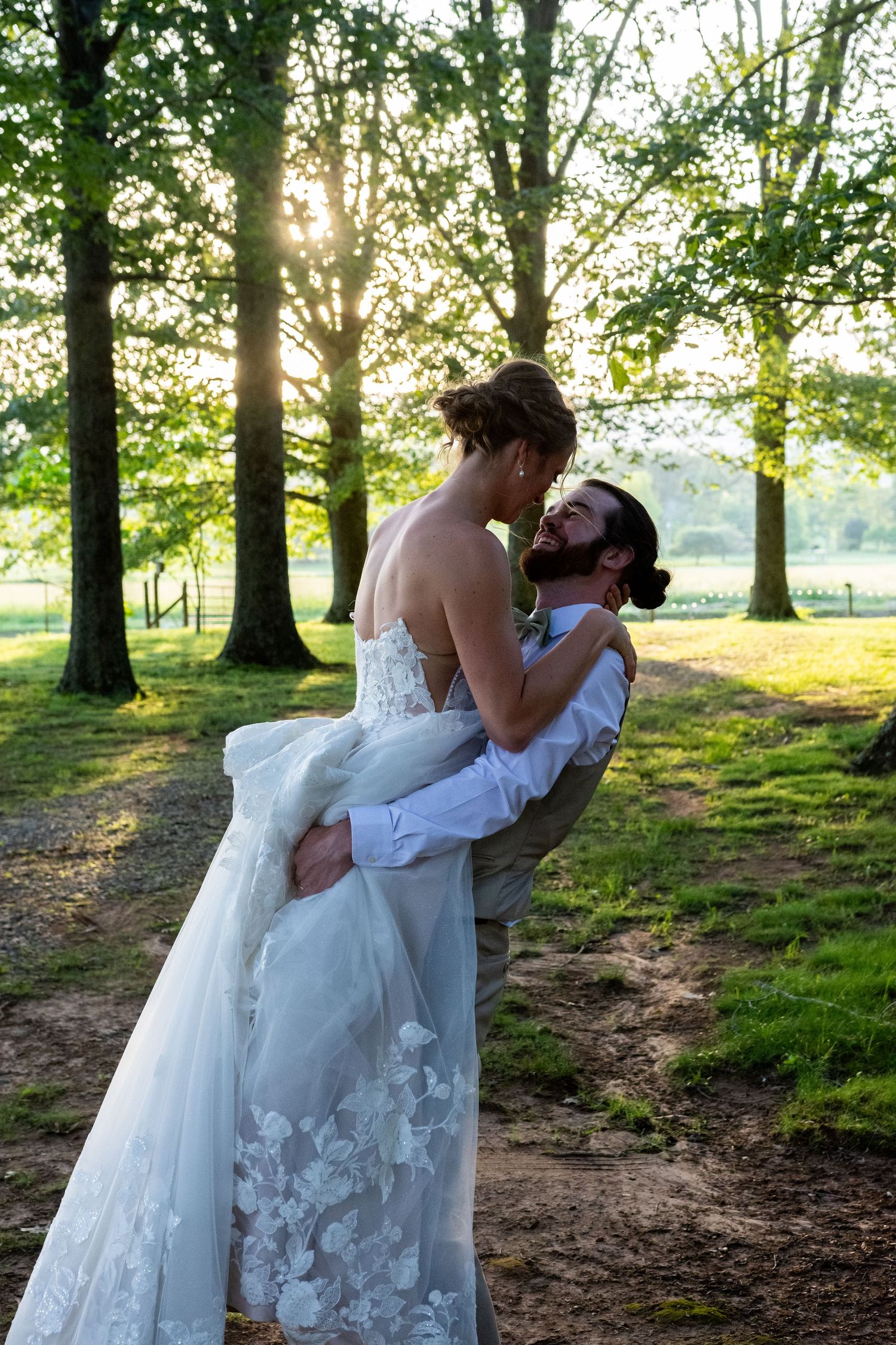 A romantic wedding couple embraces on a tree-lined path at sunset with sunlight streaming through the leaves.