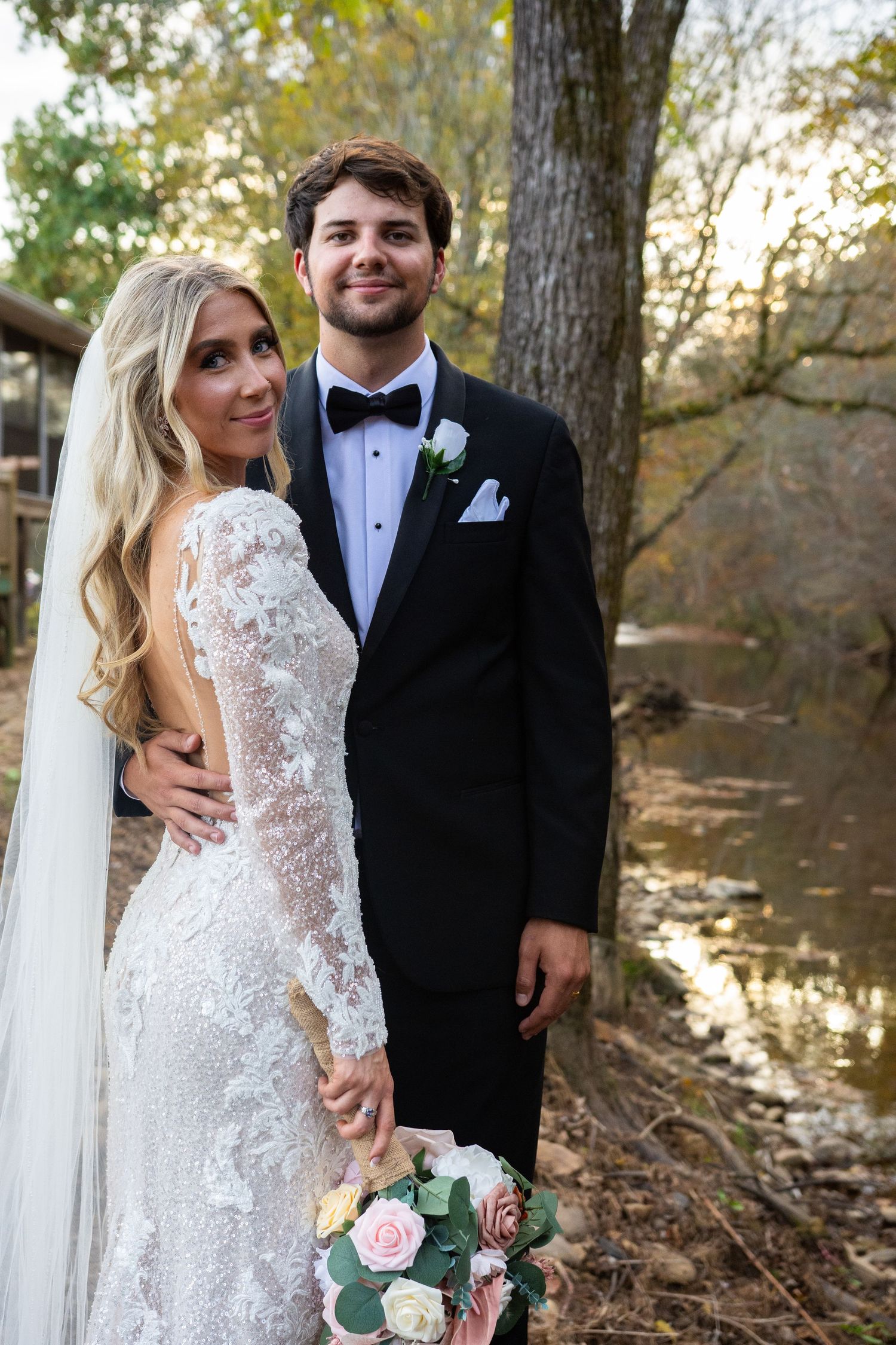 Wedding couple poses in elegant attire with white floral bouquet beside a creek during sunset at outdoor venue.