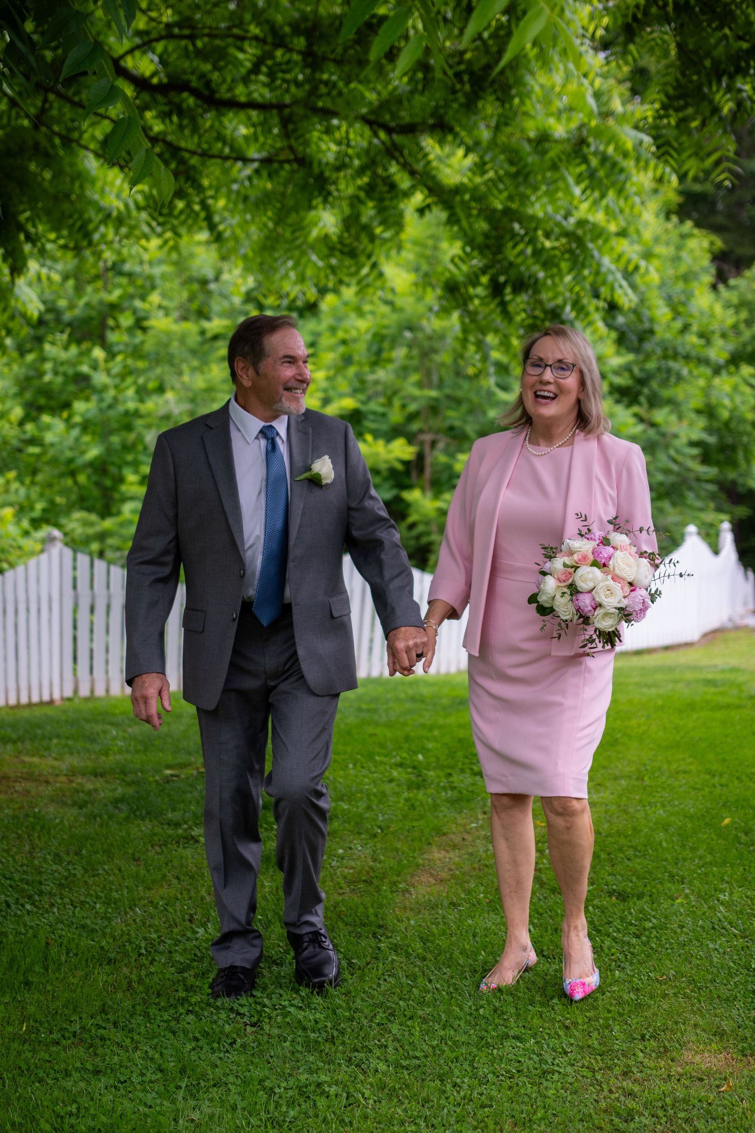 A couple walking together on green lawn, with woman in pink dress holding bouquet and man in grey suit with blue tie.
