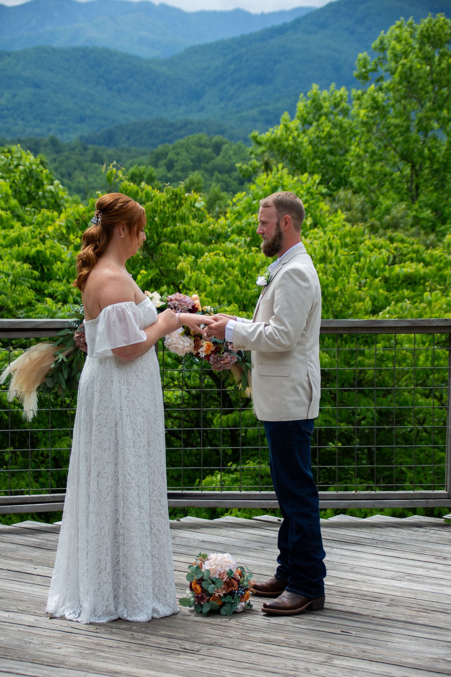 A couple exchanges wedding vows on a wooden deck overlooking lush green mountain views in summer.