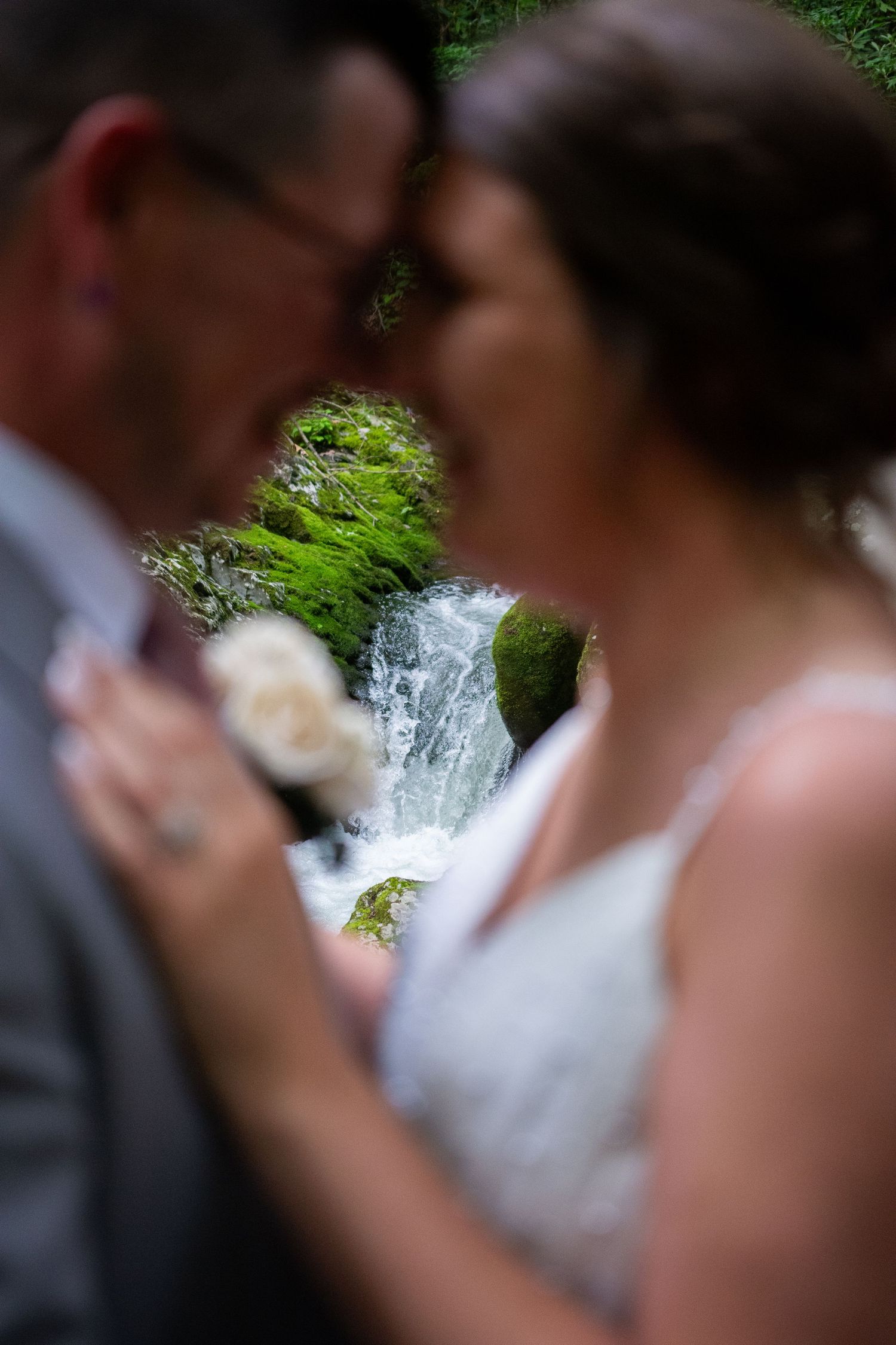 Romantic wedding couple embraces in front of scenic waterfall with lush greenery in soft focus background.