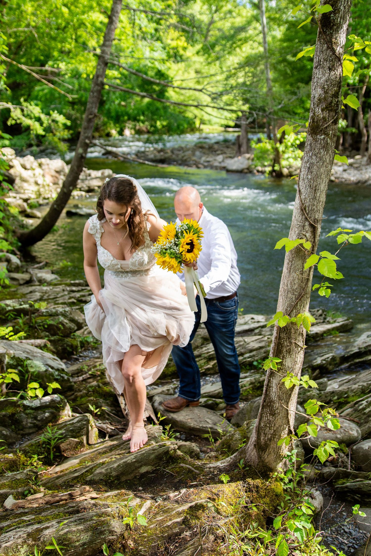 A couple in wedding attire poses together on rocky terrain by a flowing stream in a lush forest setting.