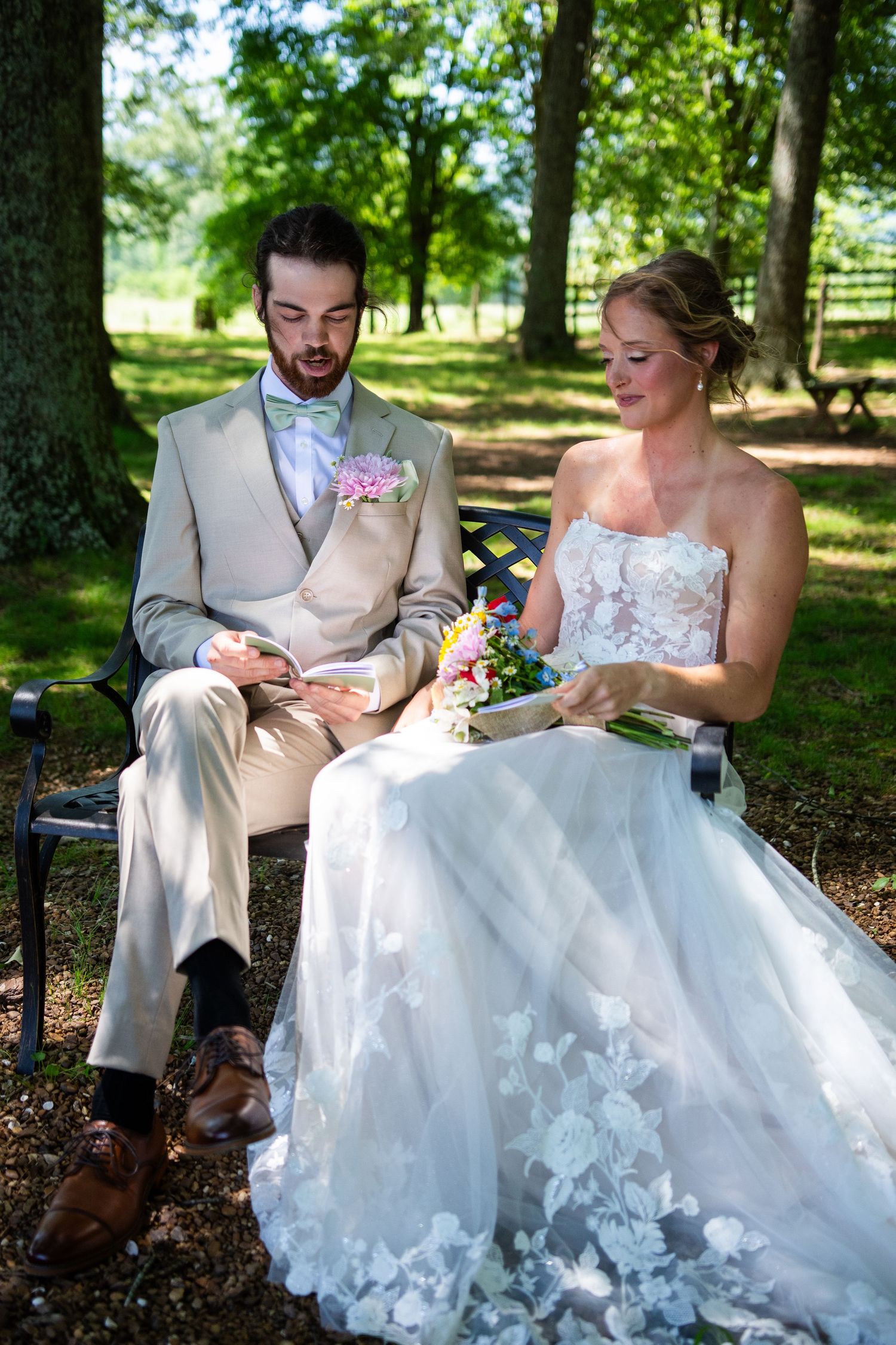 A couple sitting on a park bench sharing a quiet moment in formal attire, surrounded by lush greenery on a sunny day.
