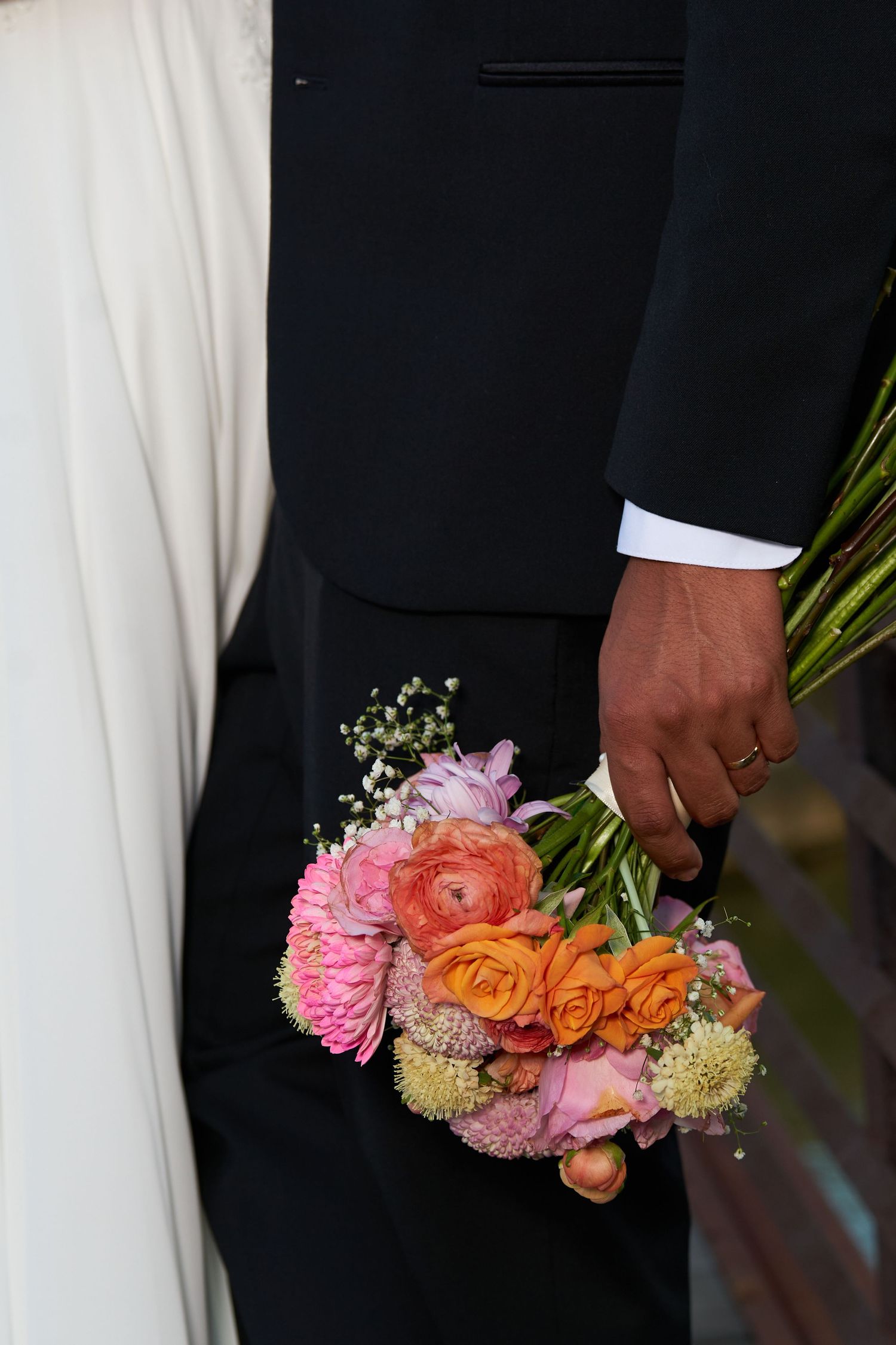 Hand in a black suit sleeve holding a colorful bridal bouquet with roses, ranunculus and baby's breath flowers.