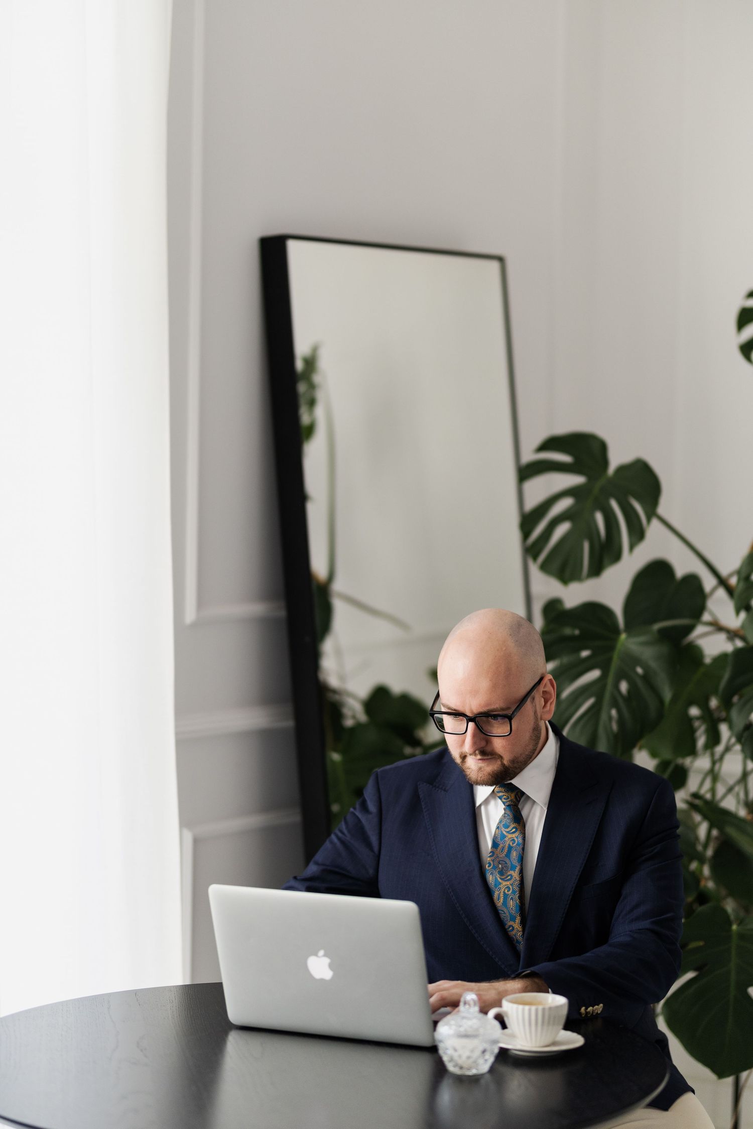 Professional works on laptop at desk with monstera plant and mirror in background.