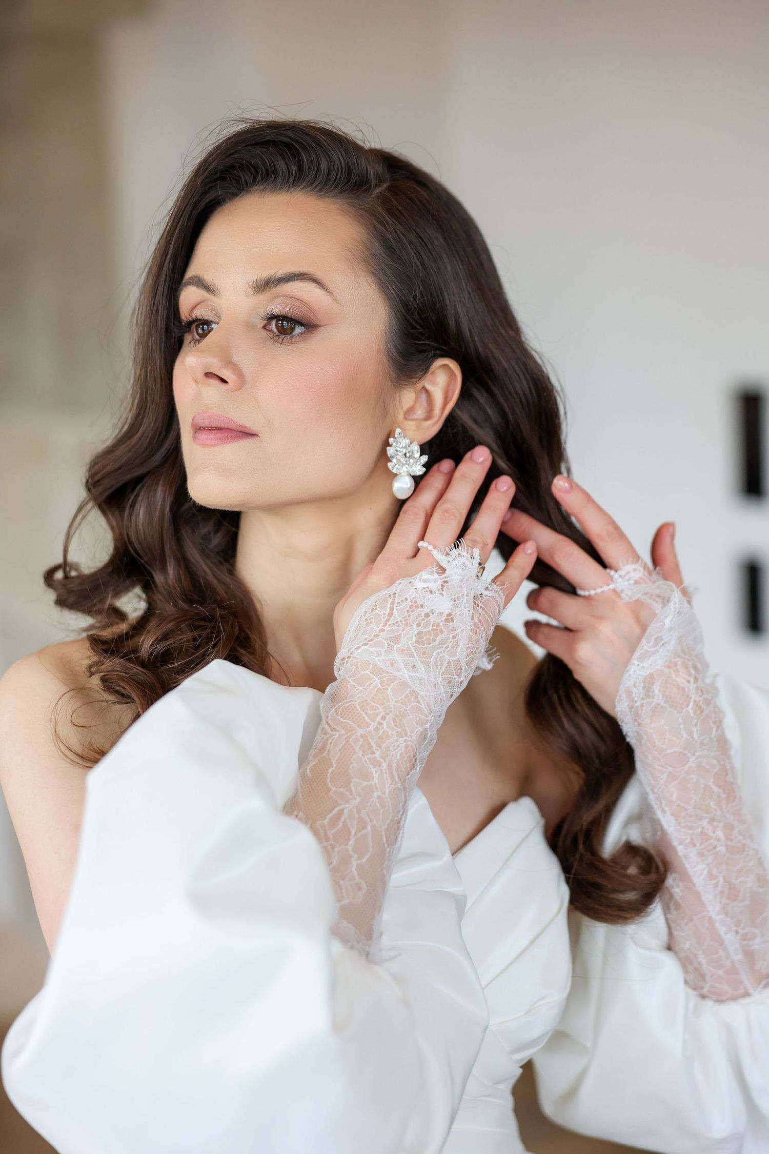 Elegant bride in white lace gloves and flowing curls adjusts her statement earring.