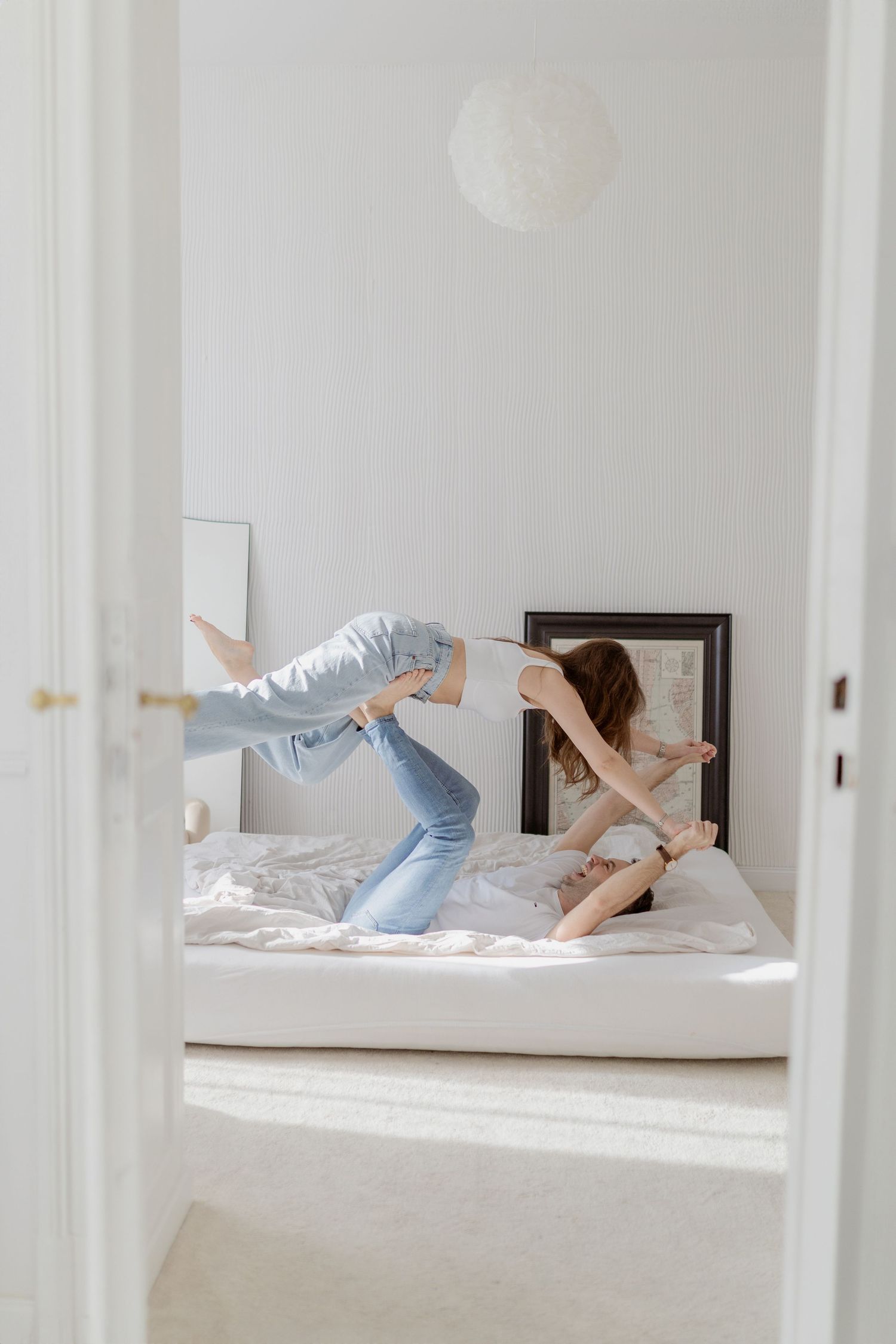 Playful scene through doorway of person jumping on white bed in bright room.