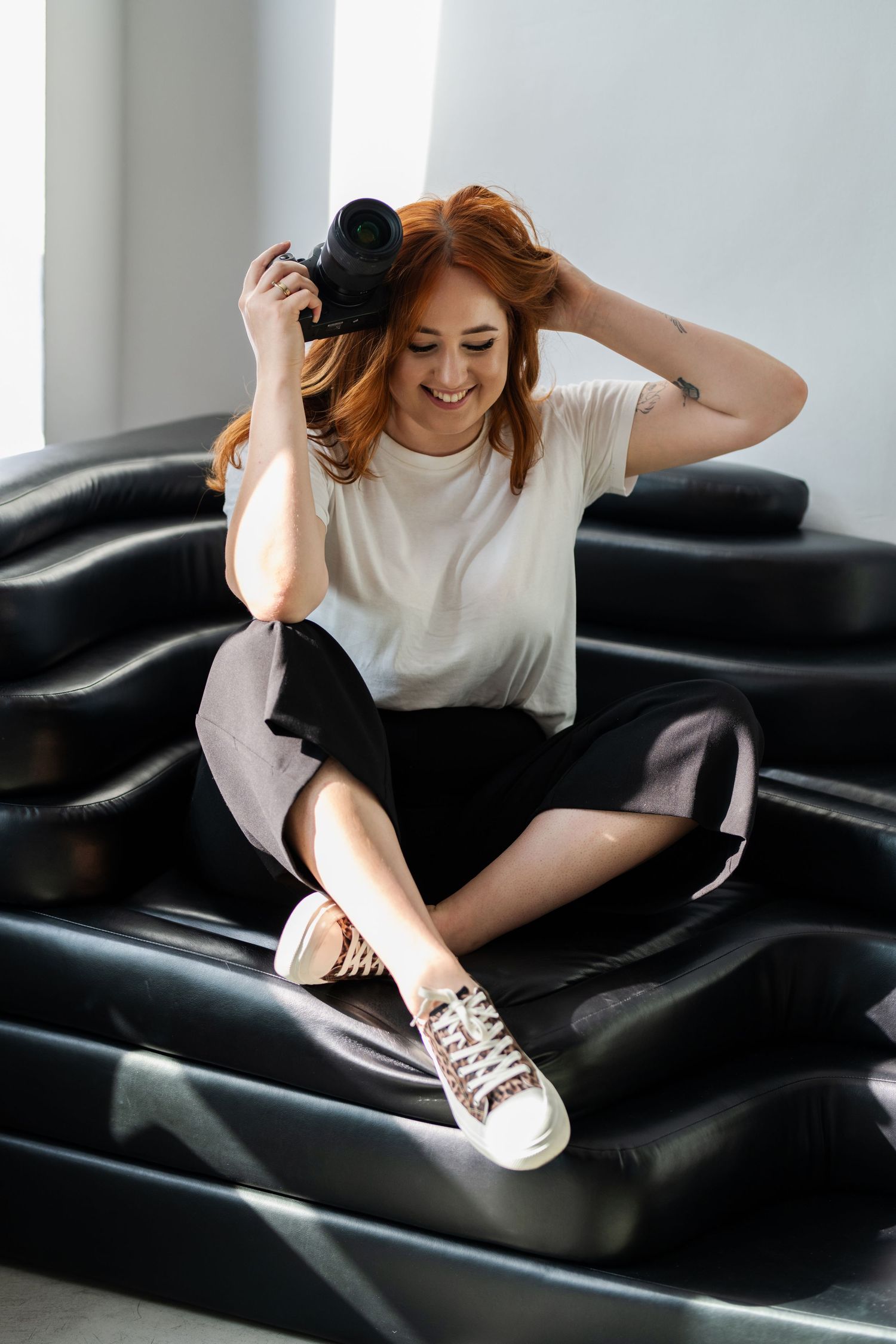 Joyful photographer with red hair holding camera while sitting on black leather furniture.
