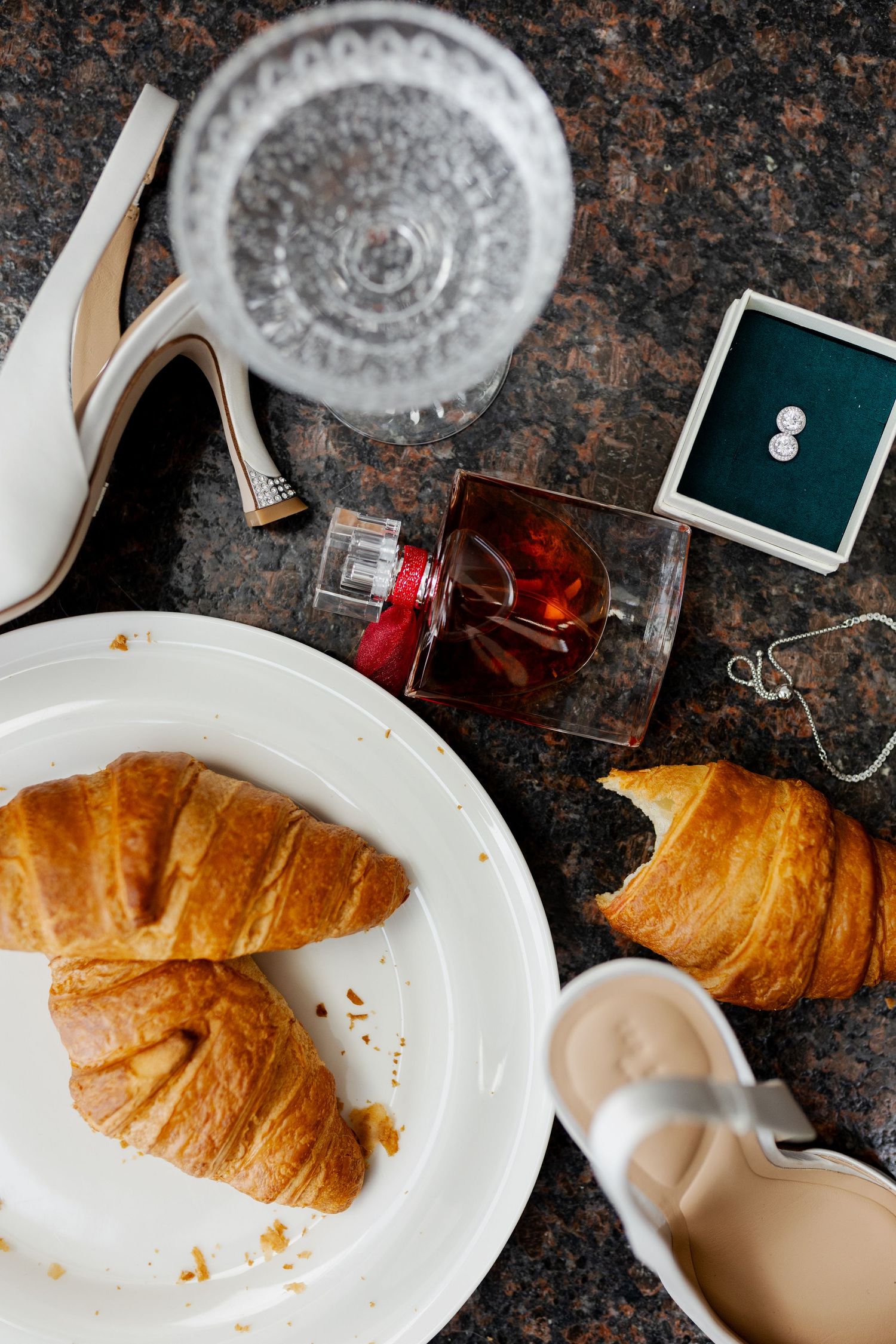 Flatlay of croissants, wedding rings, and white shoes on granite countertop.