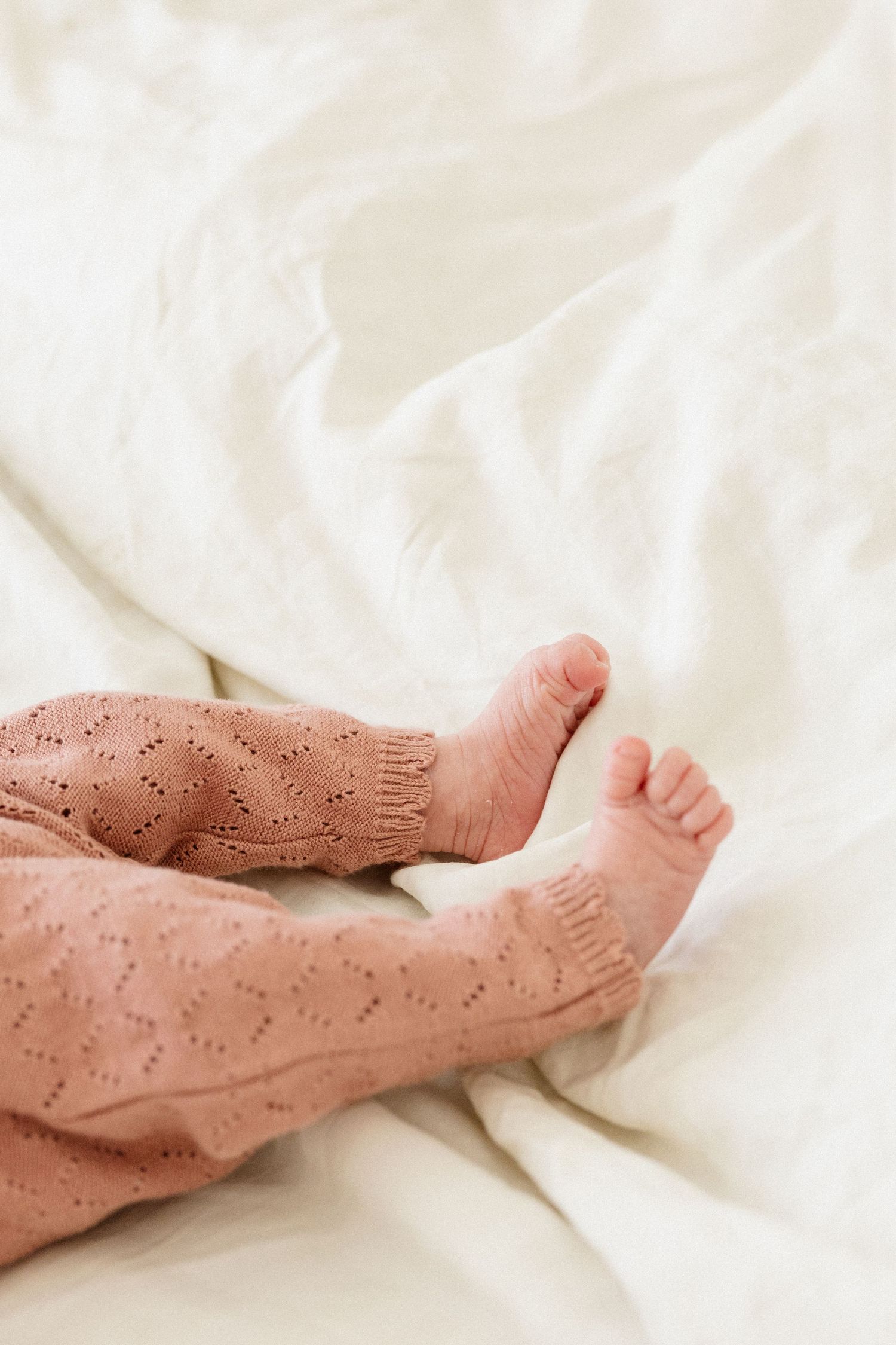 Tiny baby feet wearing knitted salmon pink leggings with heart pattern against white bedding.