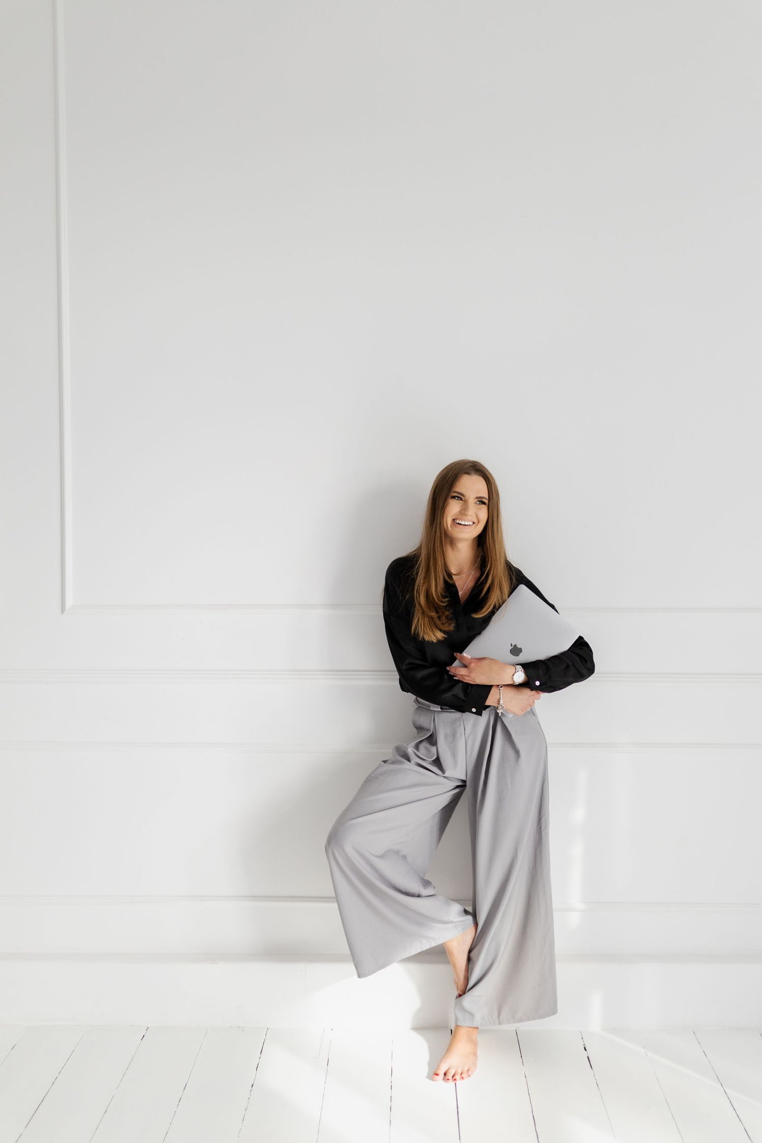 Woman in black top and flowing gray pants poses against white wall in natural light.