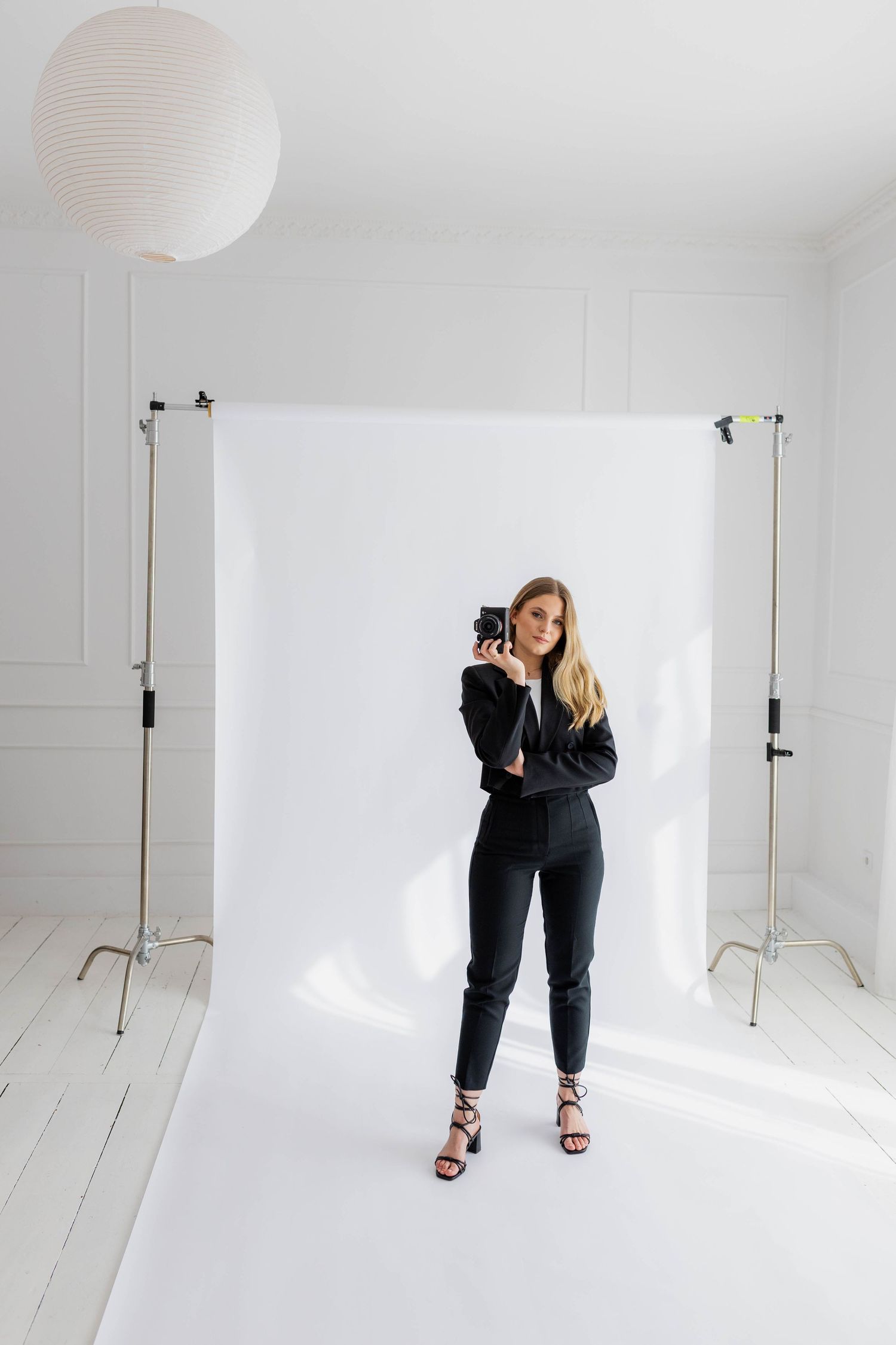 Model in black outfit poses against white backdrop in photography studio with lighting equipment.