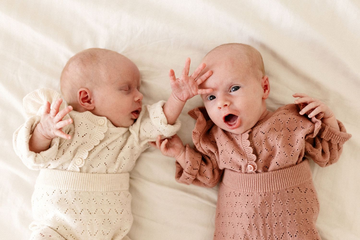 Two infants lying side by side wearing knit sweaters, one in cream and one in dusty pink, appear to be interacting.
