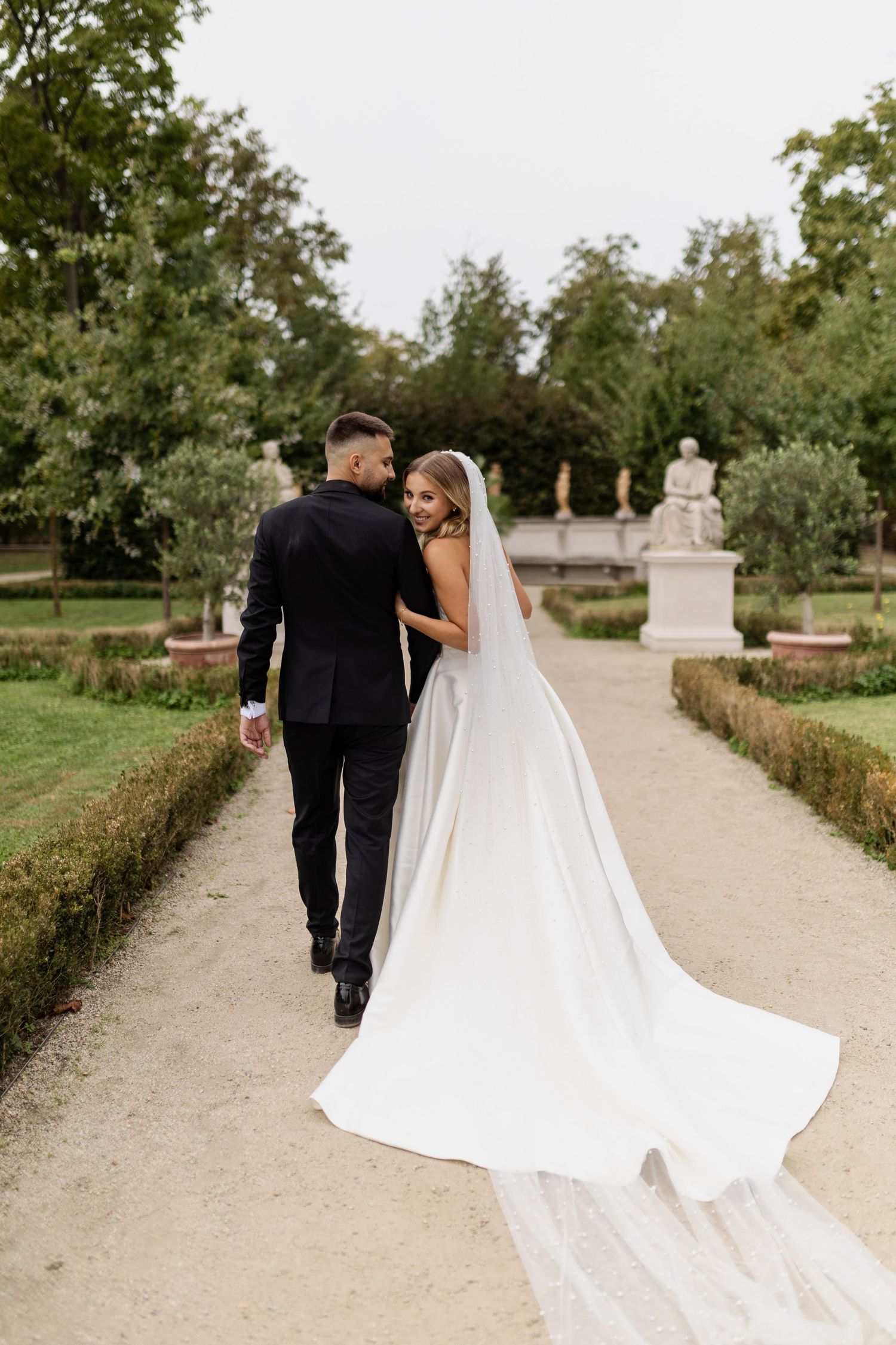 Newlyweds walk hand in hand down a garden path with trailing veil and classical statues.