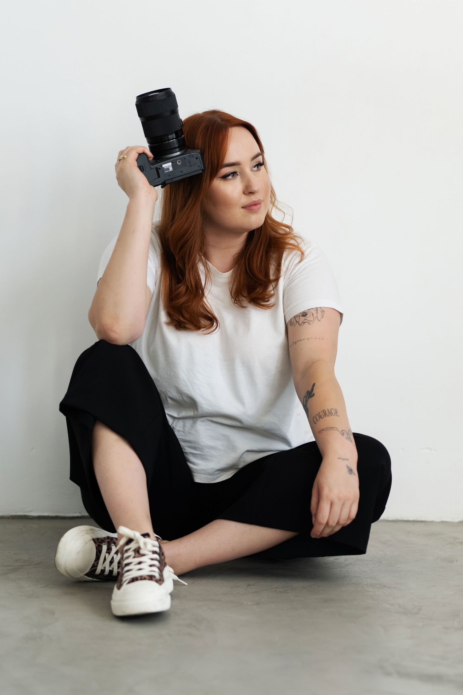 Casual portrait of photographer in white t-shirt and black pants sitting cross-legged with camera.