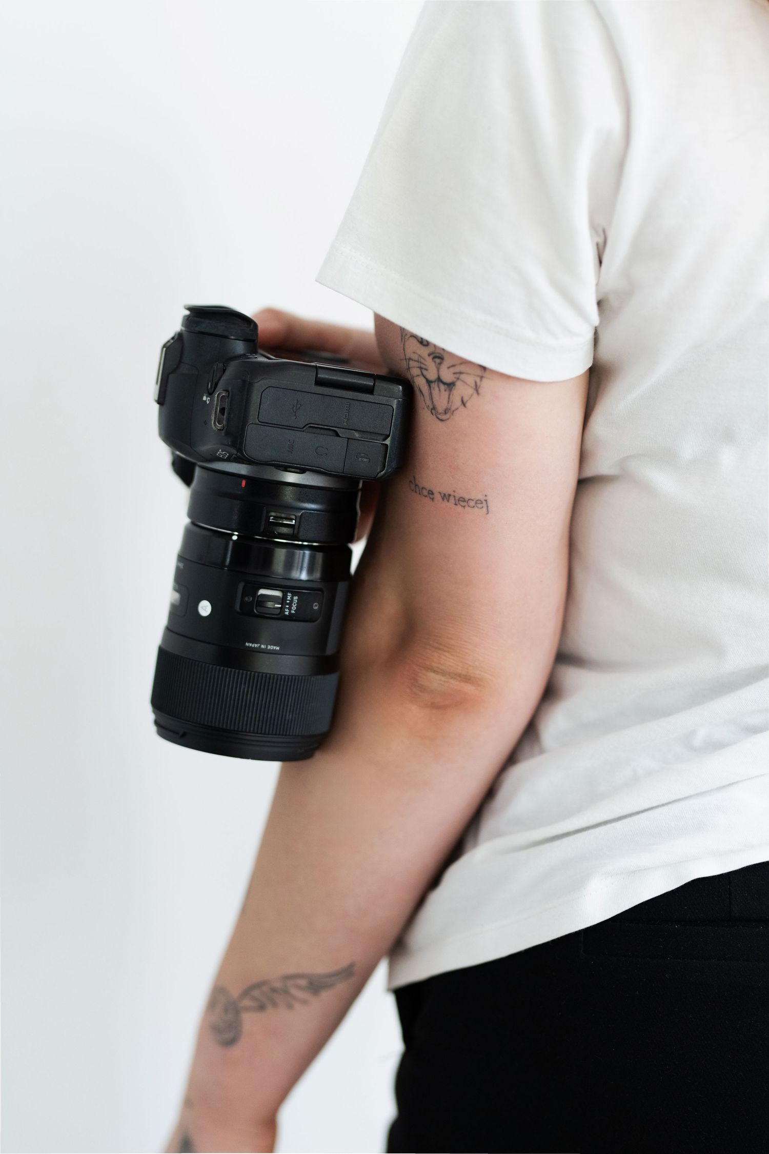 Close-up of a professional camera lens being held against a white background with visible arm tattoos.