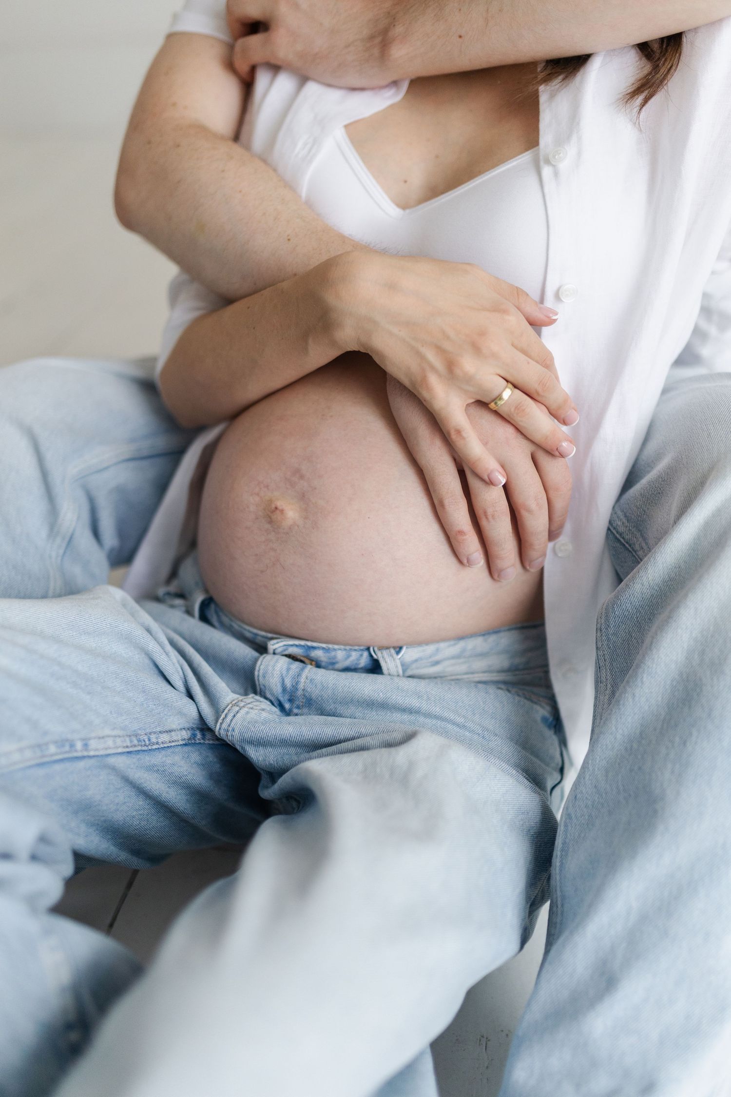 Close-up of pregnant belly with hands gently resting on it, wearing white shirt and jeans.
