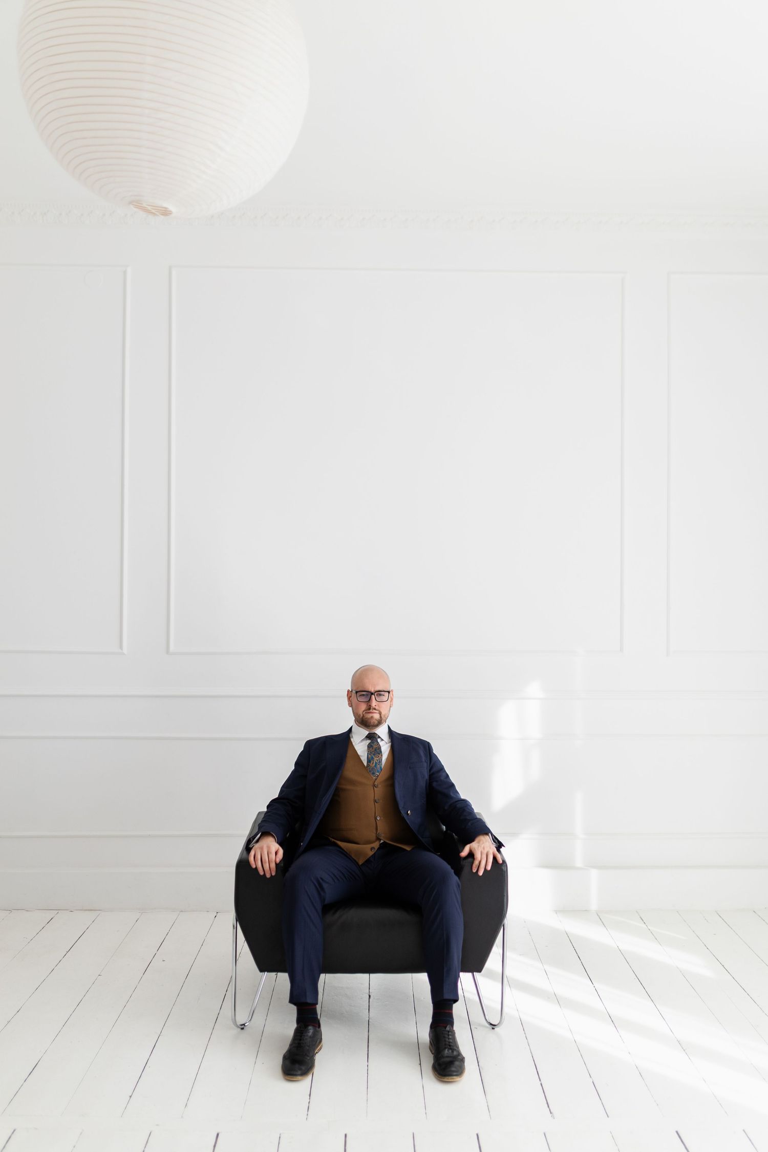 Professional in navy suit and brown vest sits centered in minimalist white room with paper lantern.