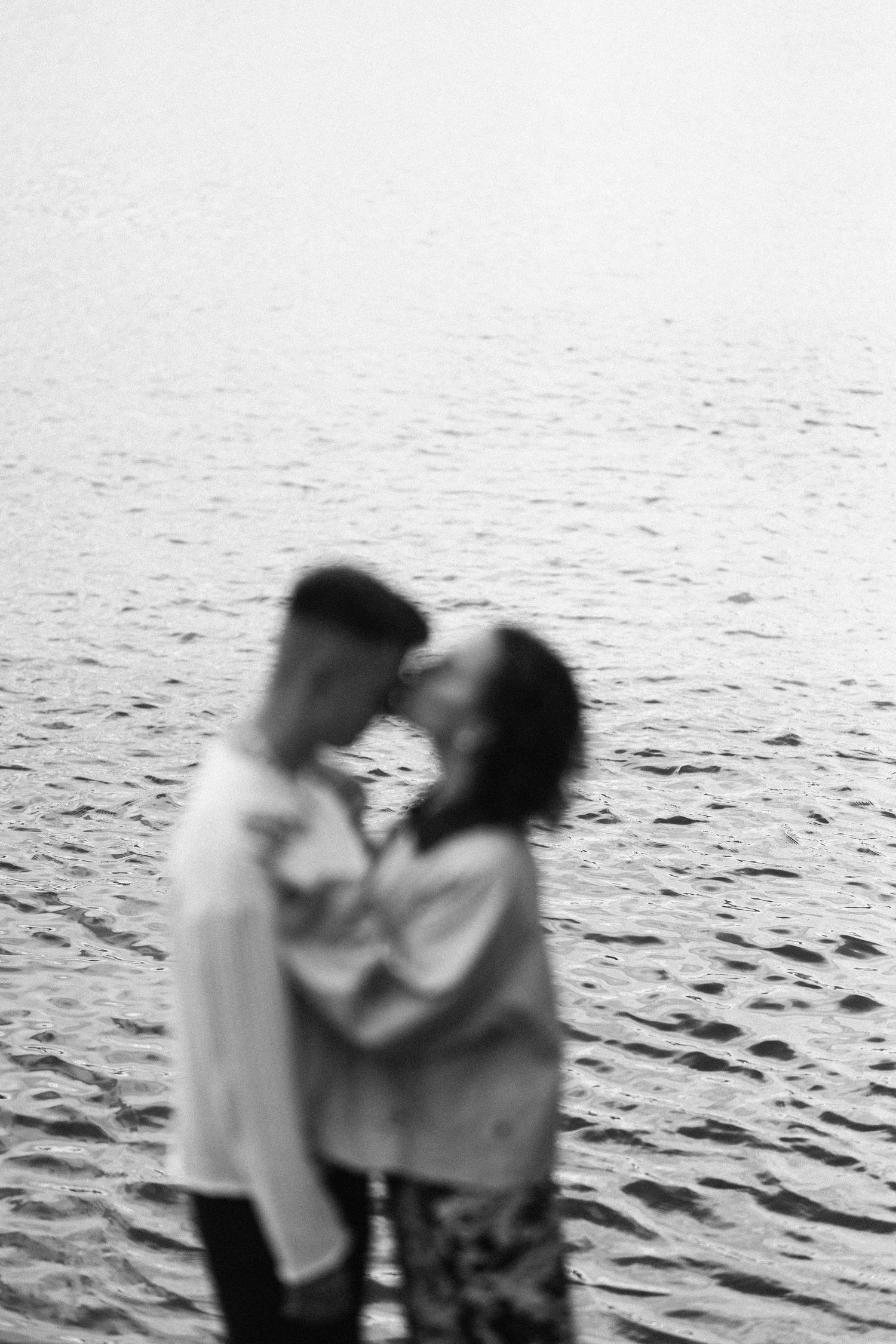 Blurry black and white silhouette of couple embracing by water at sunset.