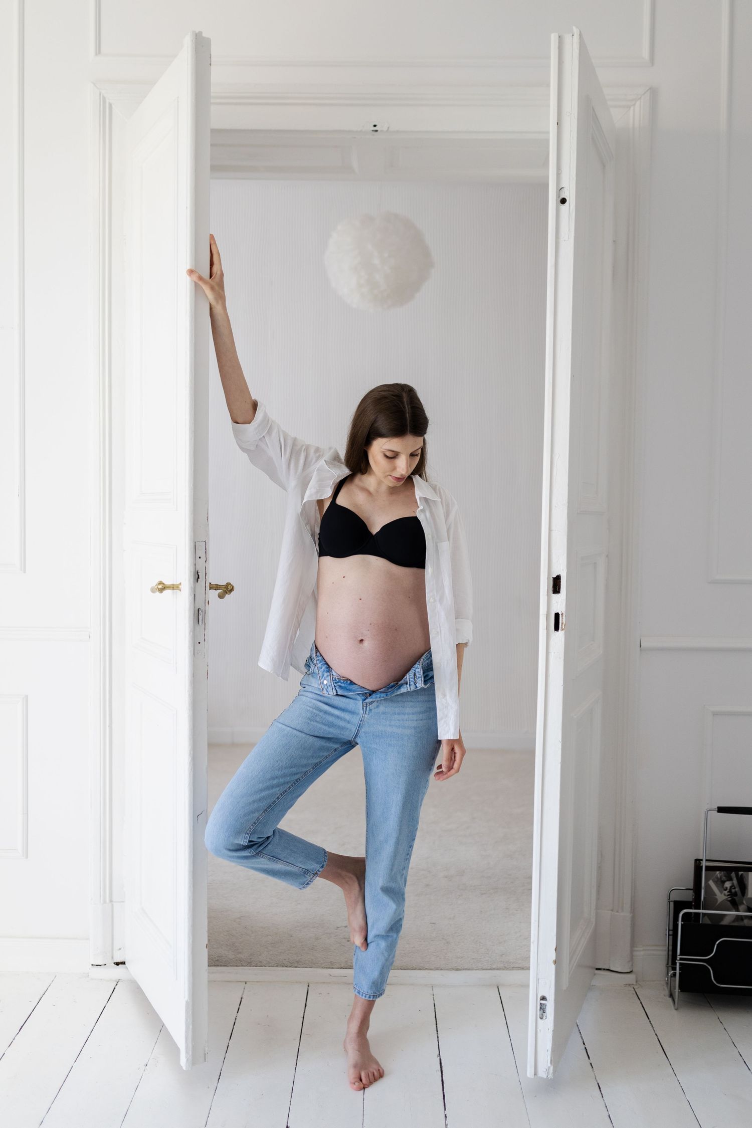 Expectant person in casual denim and white shirt strikes playful pose in white doorway with paper lantern above.