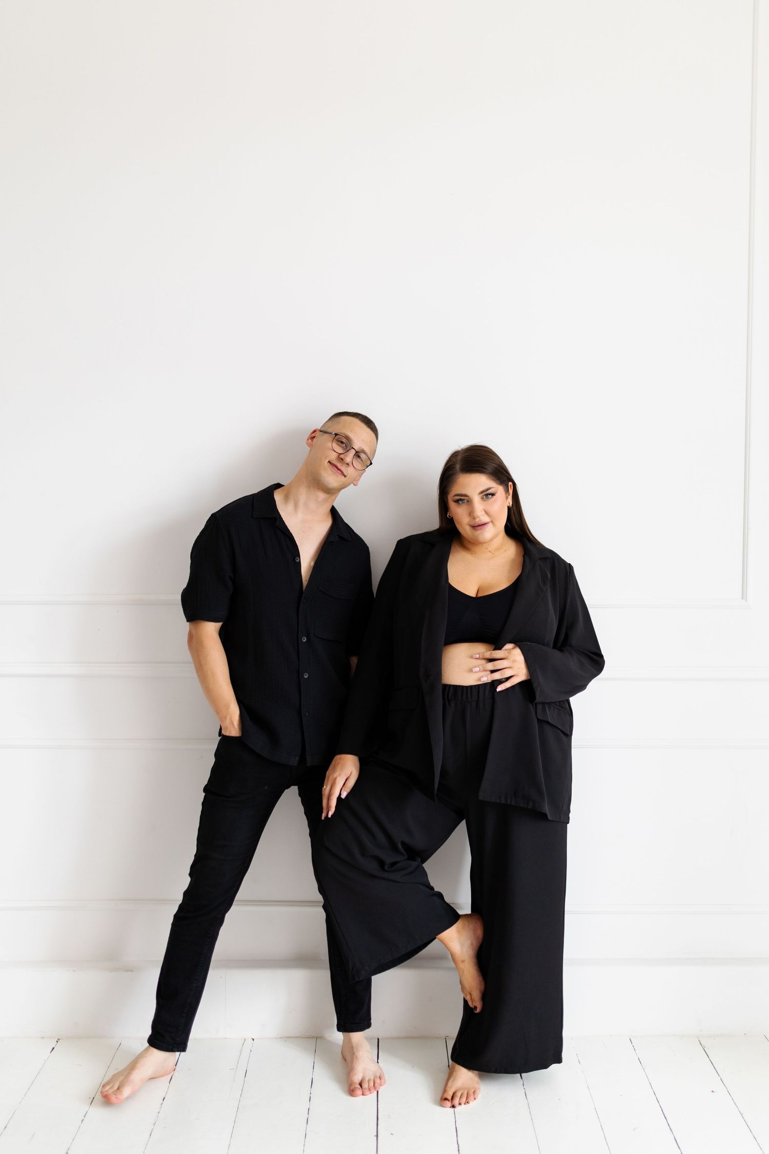 Couple in matching black outfits posing against white wall in casual lifestyle portrait.