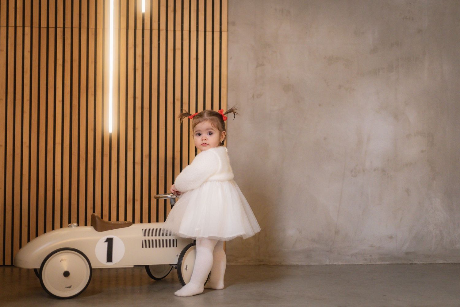 A toddler in a white dress stands next to a vintage toy car against a wooden panel wall.