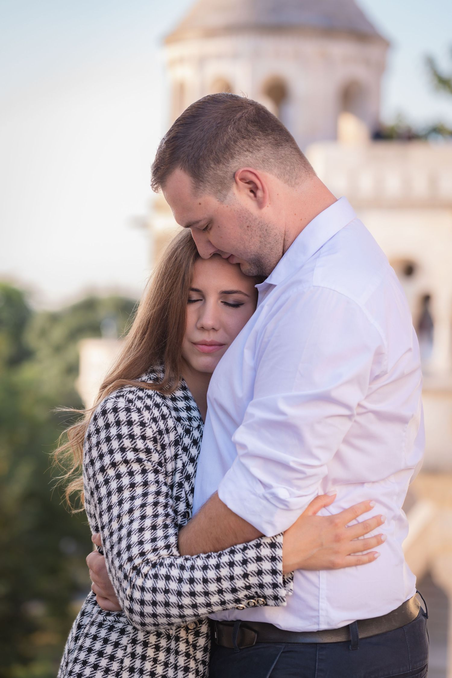 Tender moment captured between couple in checkered dress and white shirt near historic building.