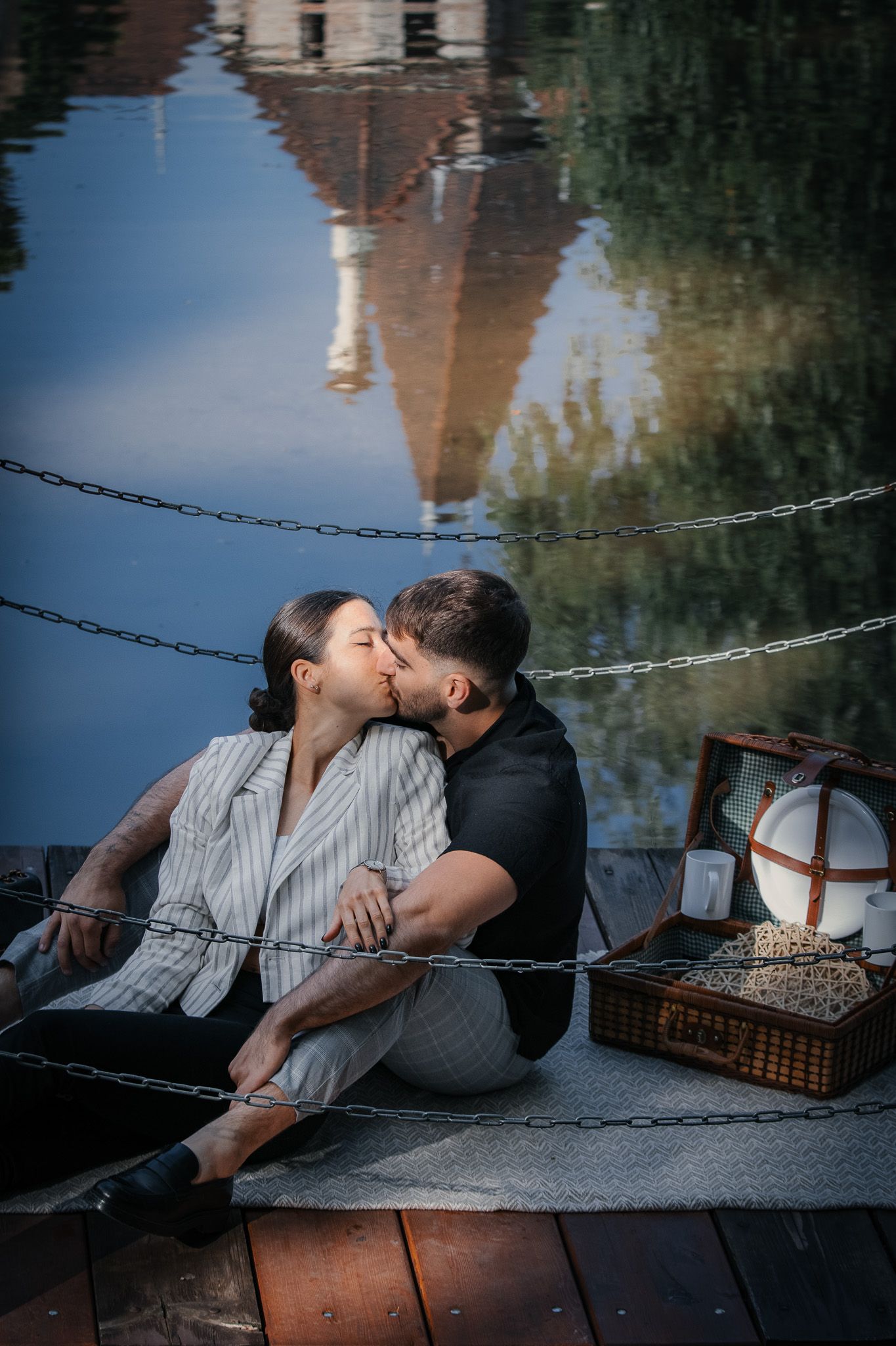 Romantic picnic scene by waterfront with couple relaxing on blanket near reflective water.