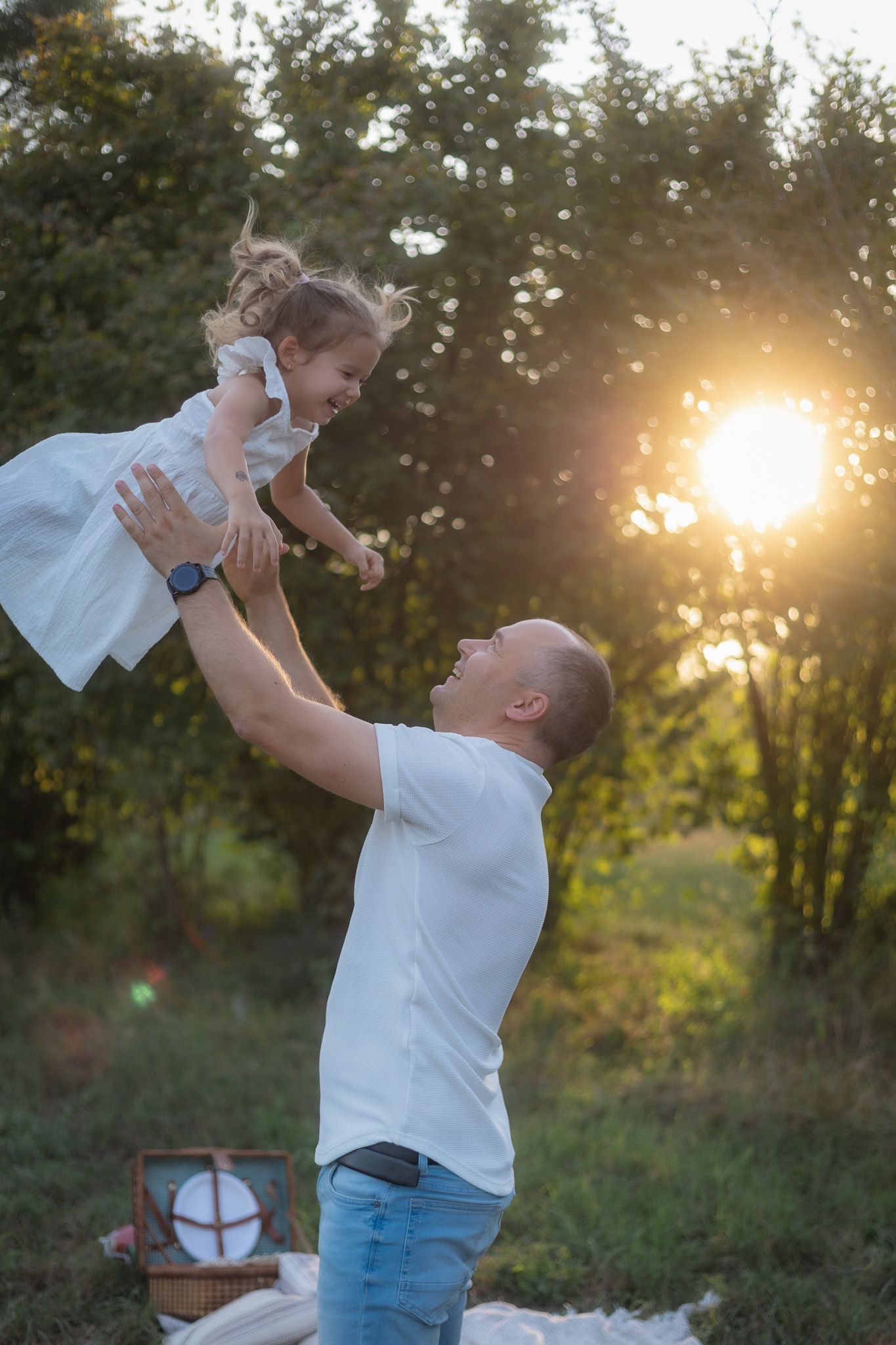 Person in white shirt lifting child in white dress during sunset outdoor photoshoot.