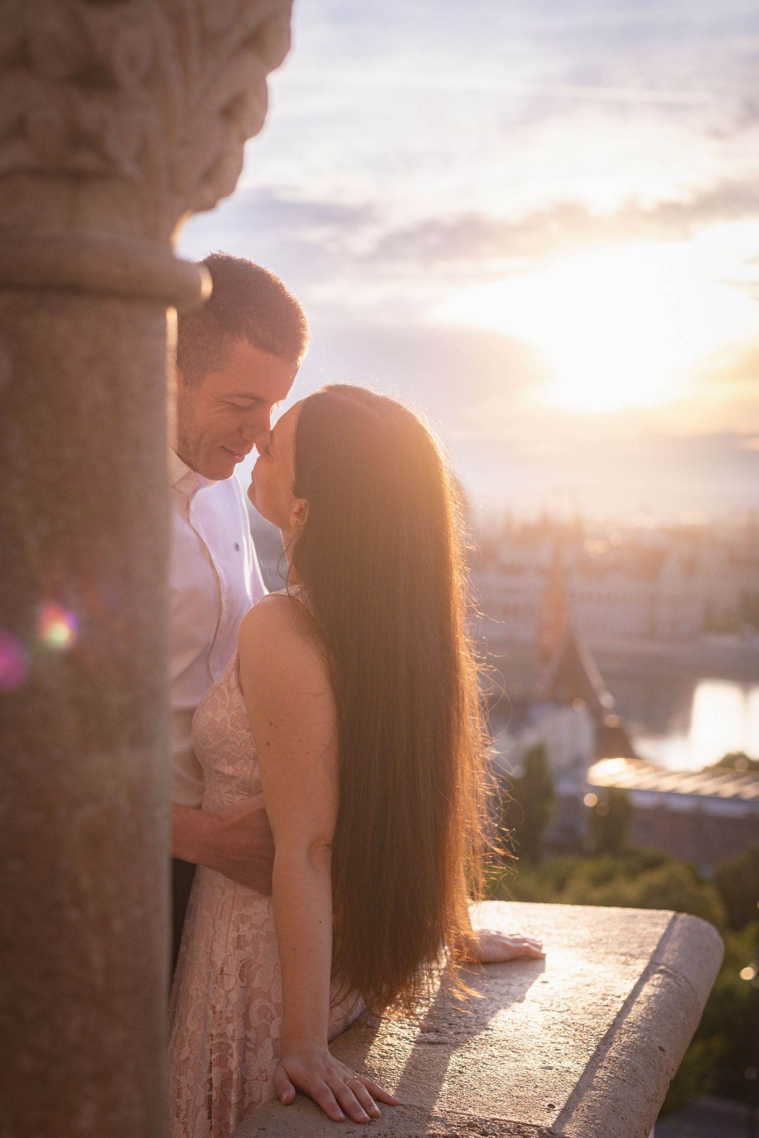 A couple shares an intimate moment at sunset on a stone balcony overlooking an urban skyline.