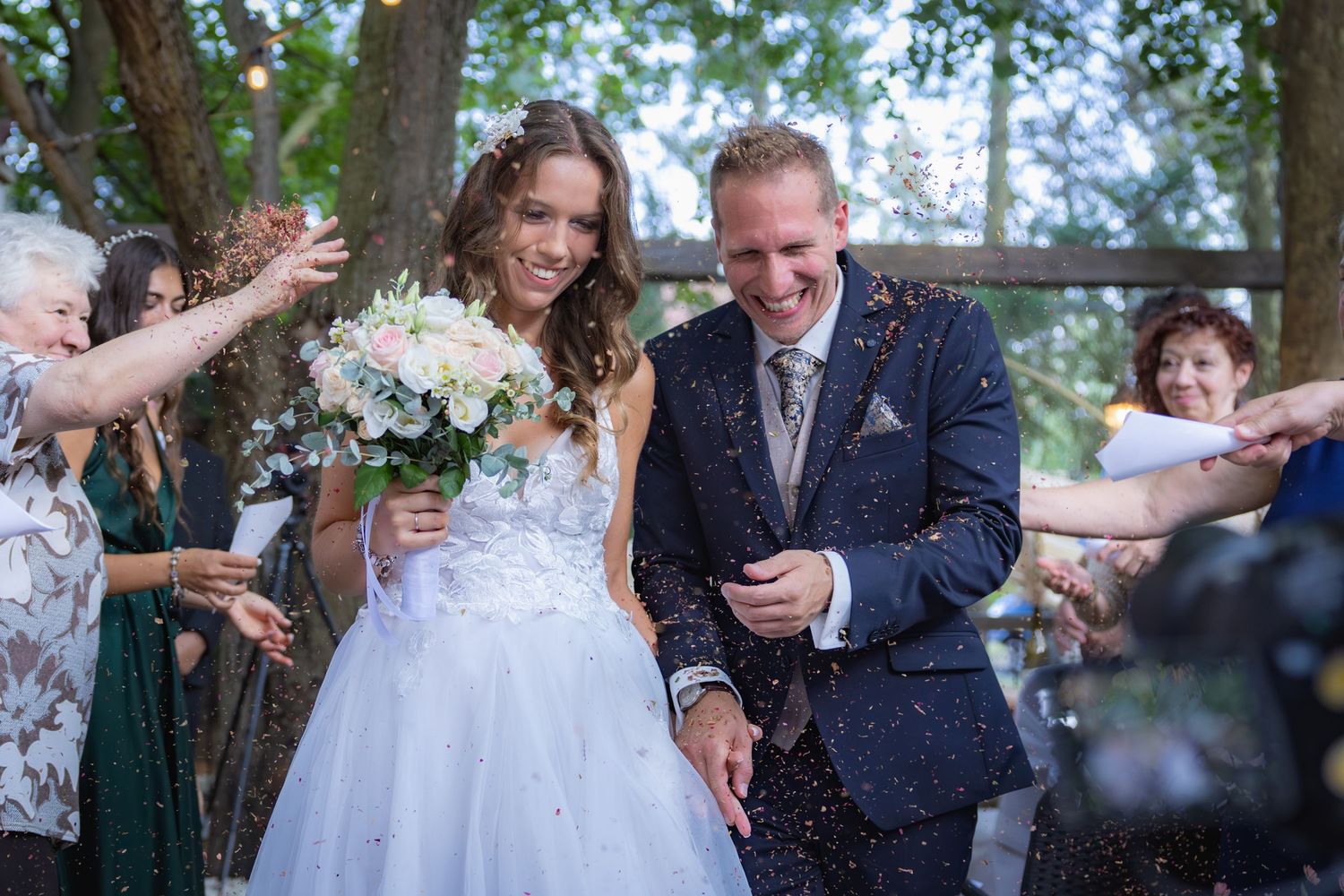 Joyful wedding couple walking together outdoors while guests throw flower petals and confetti in celebration.