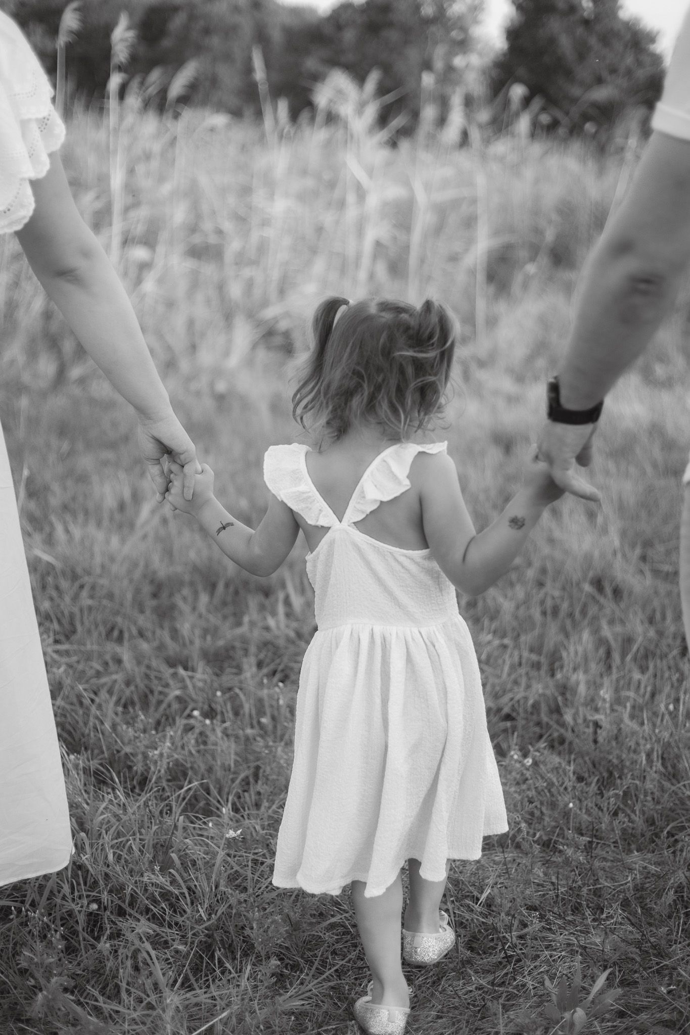 A young child in a white dress holds hands with adults while walking through a field in black and white.