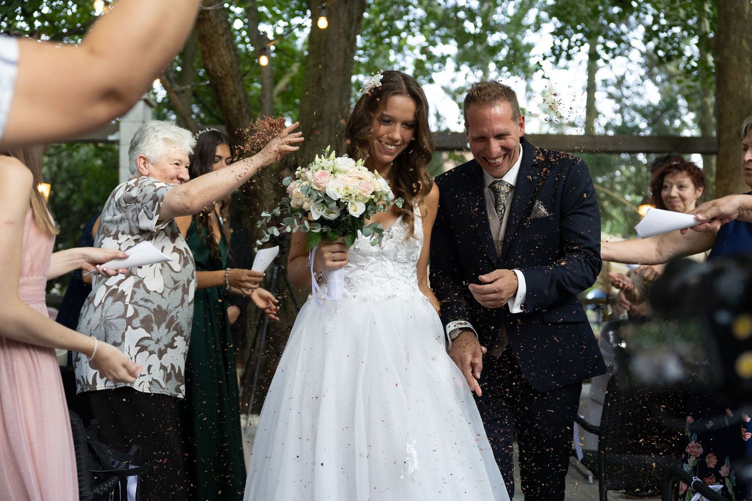 Joyful newlyweds being showered with confetti by guests during outdoor wedding celebration.