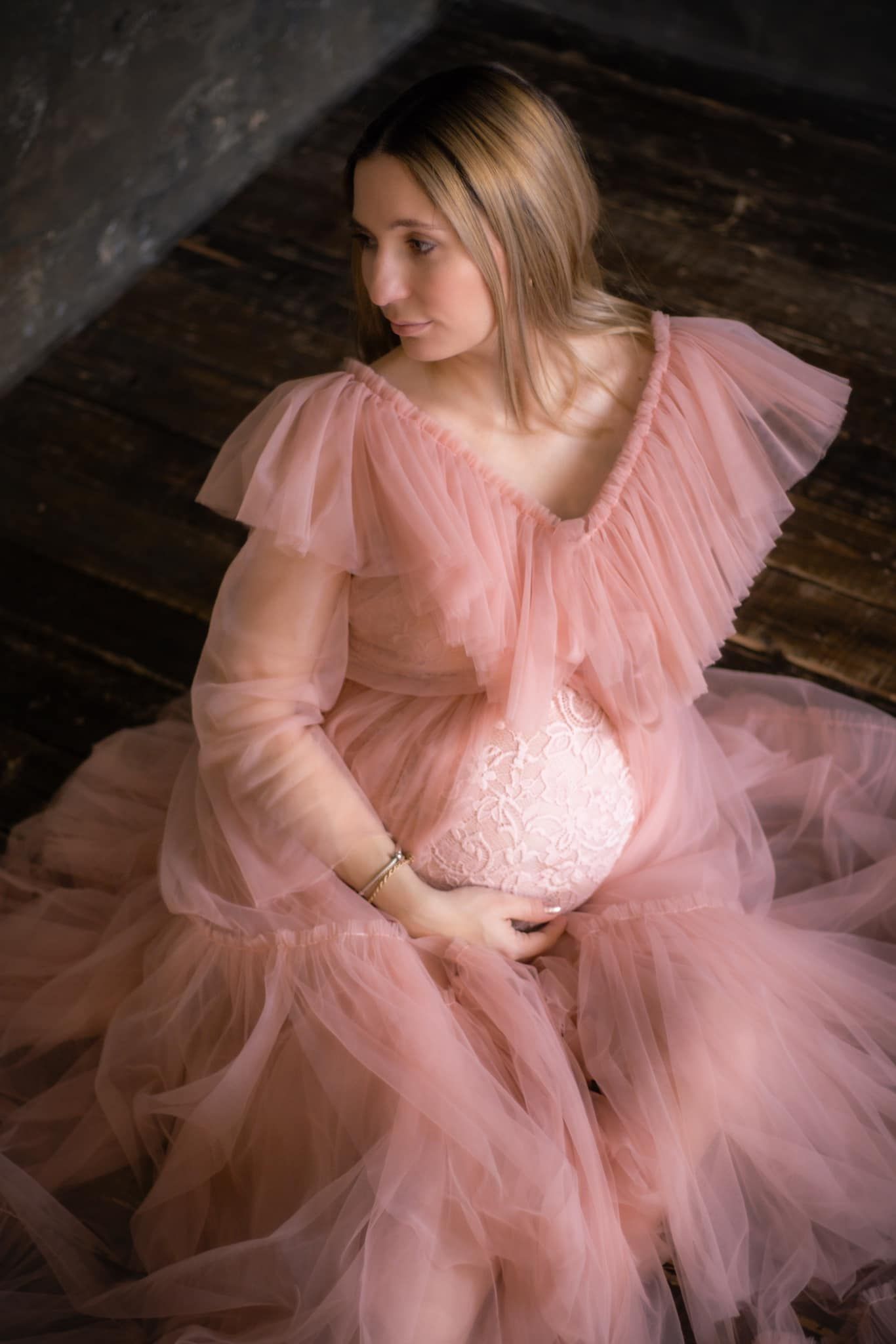 Ethereal maternity portrait featuring flowing pink tulle gown against rustic wooden floor.