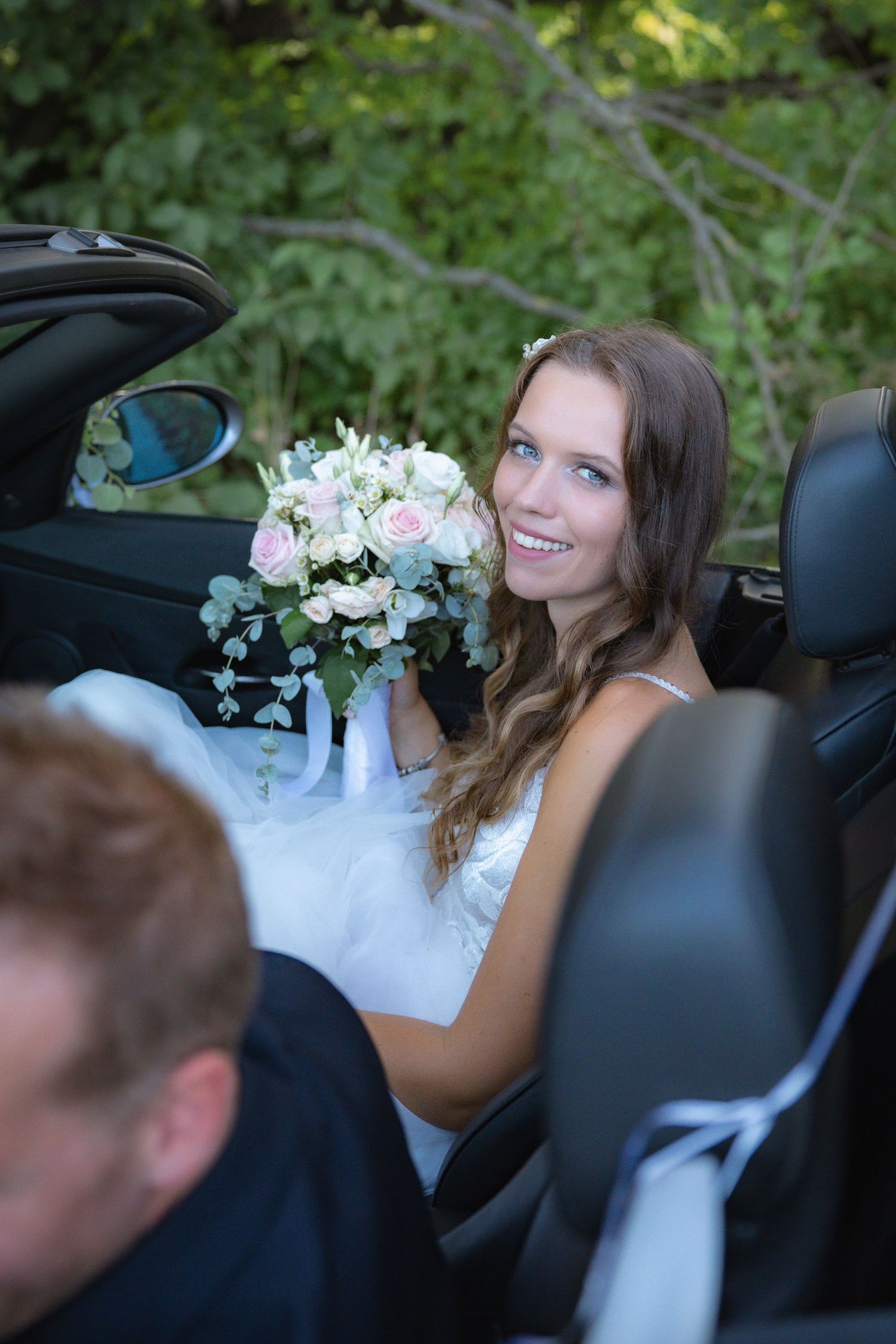 Bride sitting in convertible car holding elegant white and pink floral bouquet.
