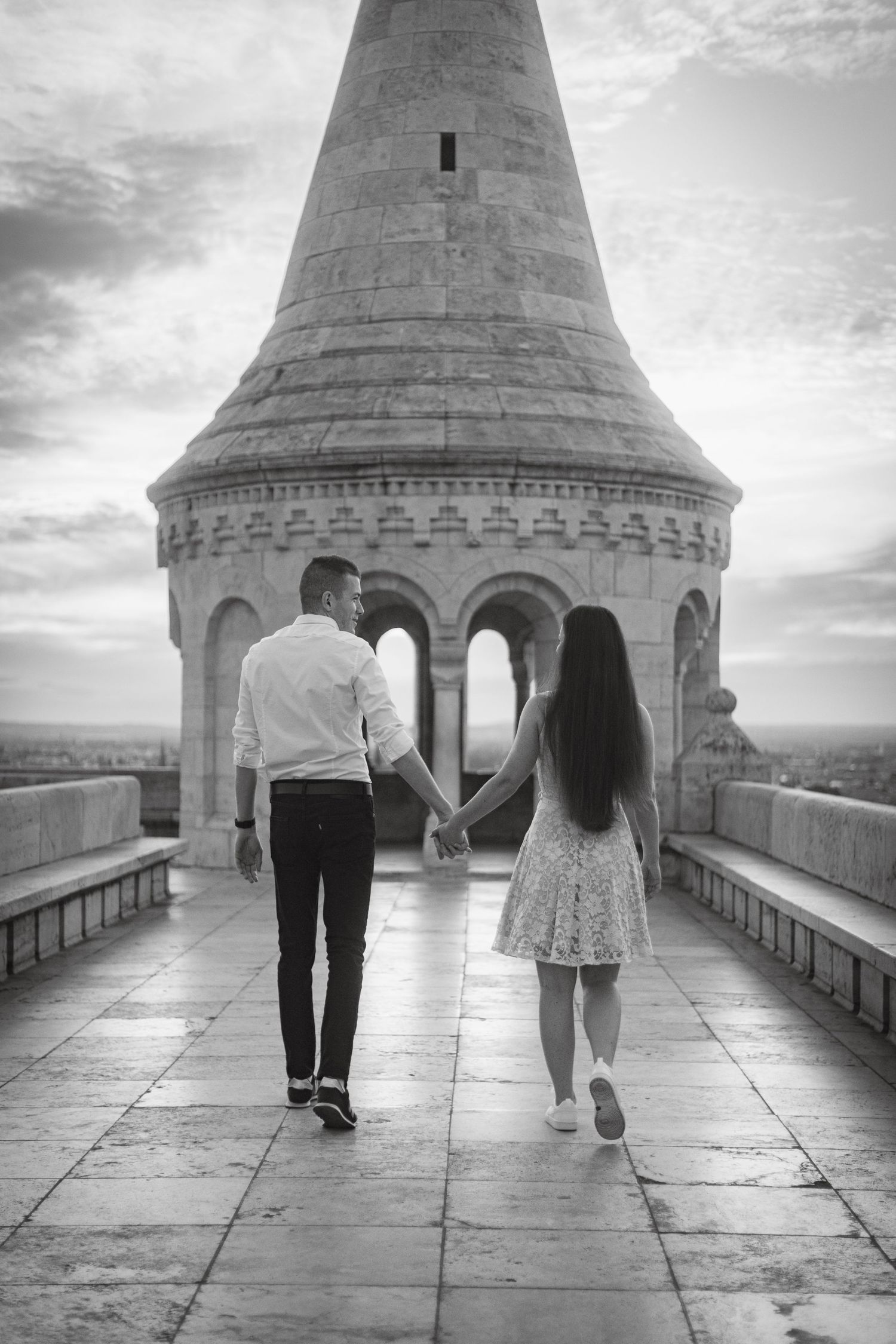 A romantic couple holds hands while walking along the stone pathway of a historic tower with arched windows and city views below.
