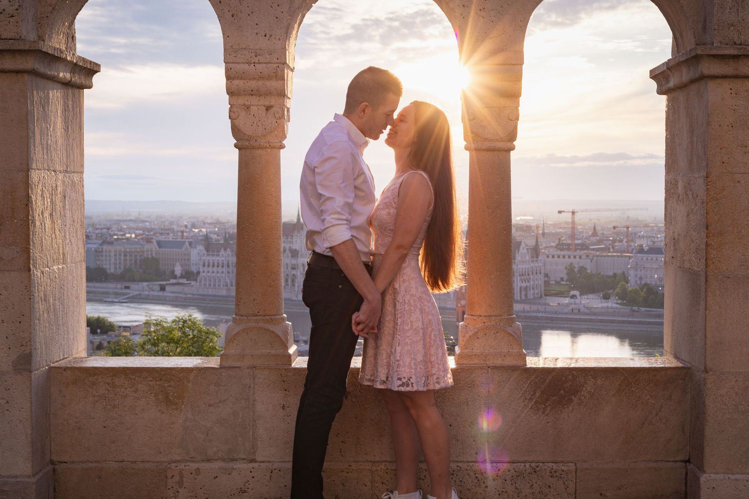 A couple shares a romantic moment in an ornate stone archway overlooking a sunset cityscape.