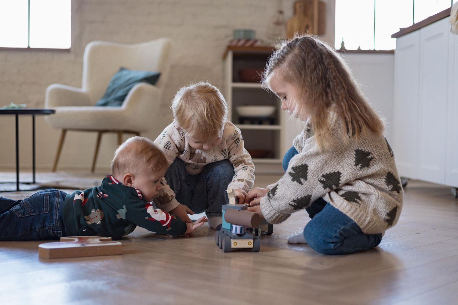 Young children play together with toys on a hardwood floor in a bright sunny room.