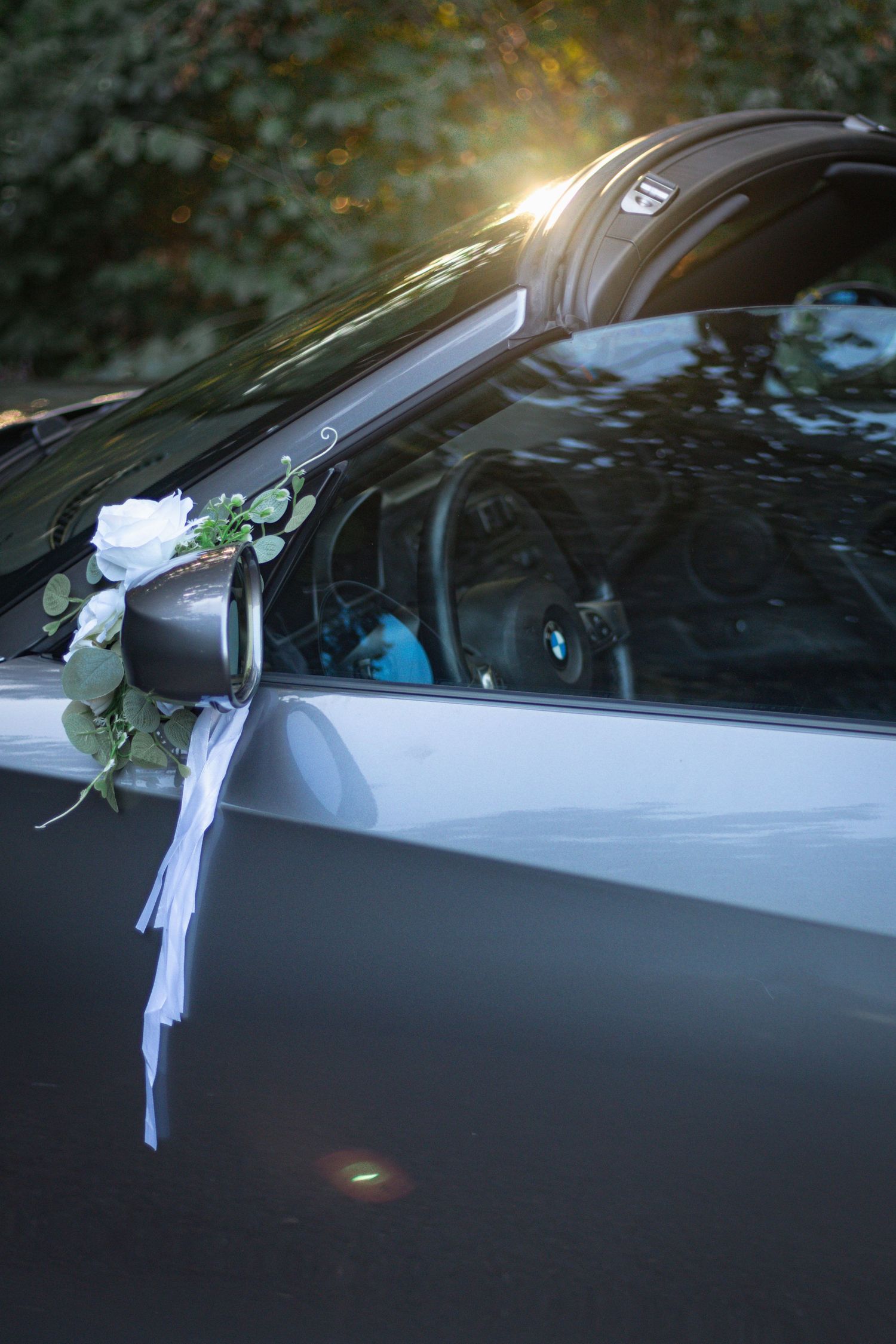 Wedding car decorations with white ribbons and flowers on a dark vehicle.