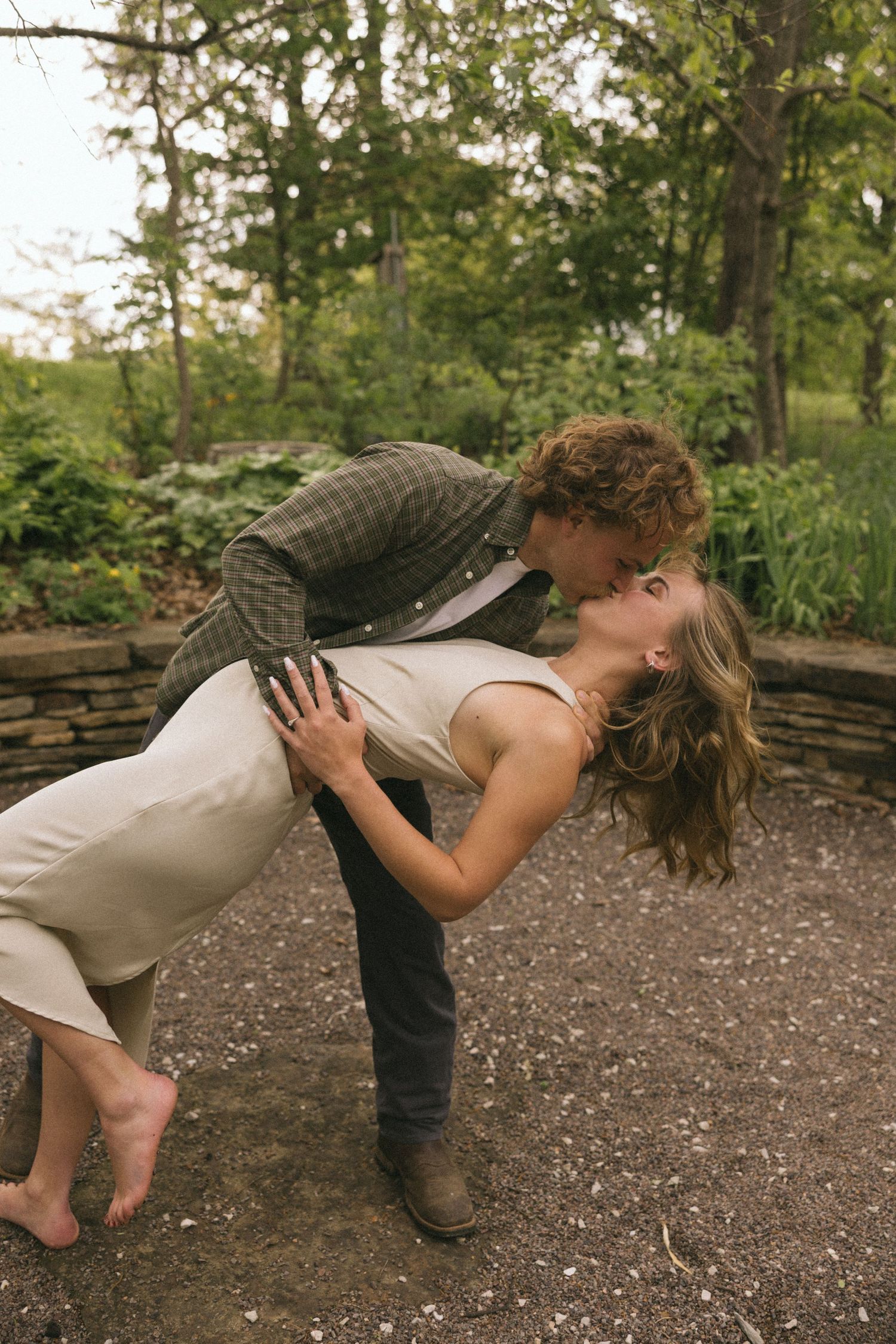 A romantic dip kiss moment captured in a garden setting with stone walls and trees in the background.