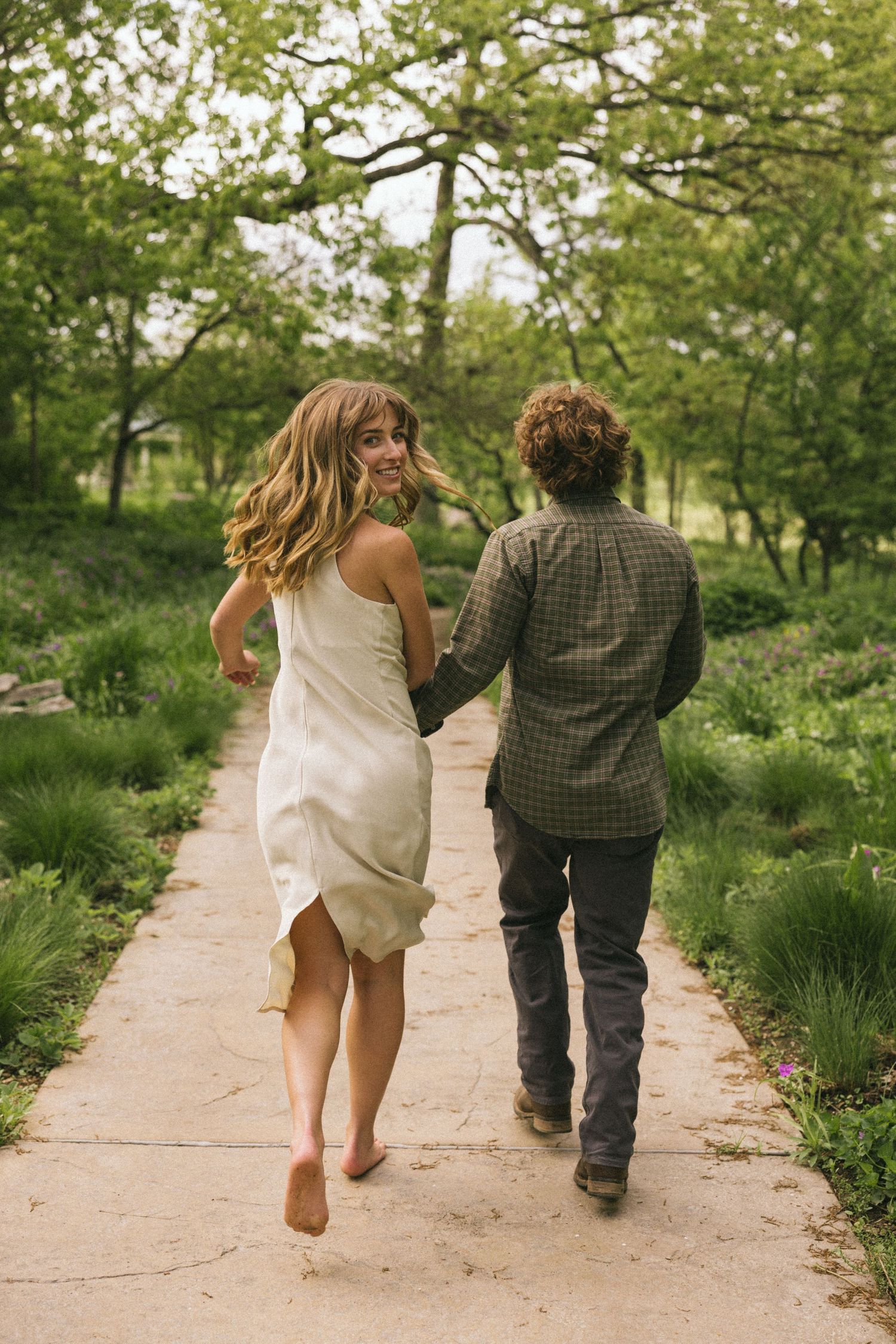 Two people hold hands while walking together down a rustic garden path surrounded by lush green foliage.