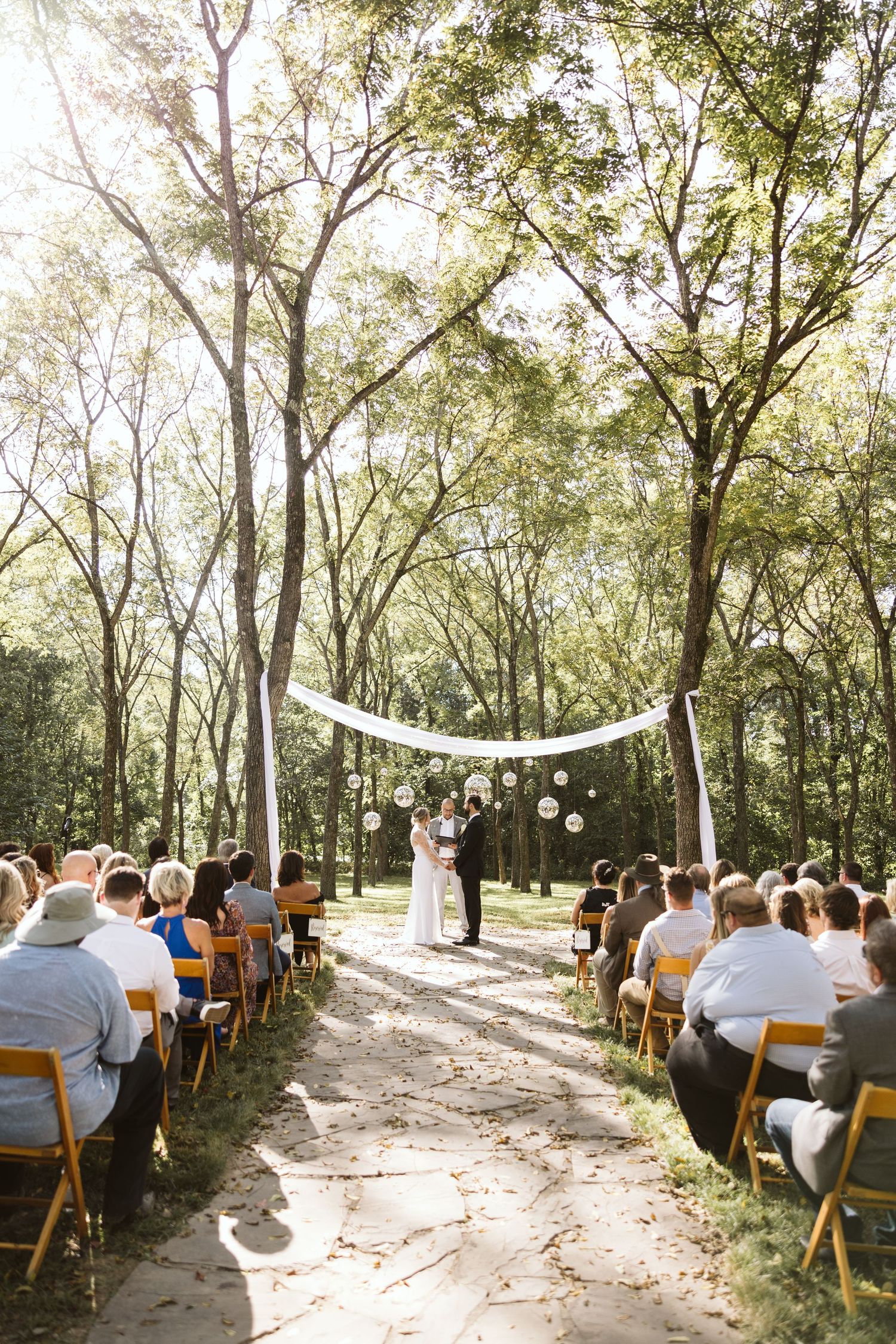 Outdoor woodland wedding ceremony setup with wooden chairs and natural sunlight filtering through trees.