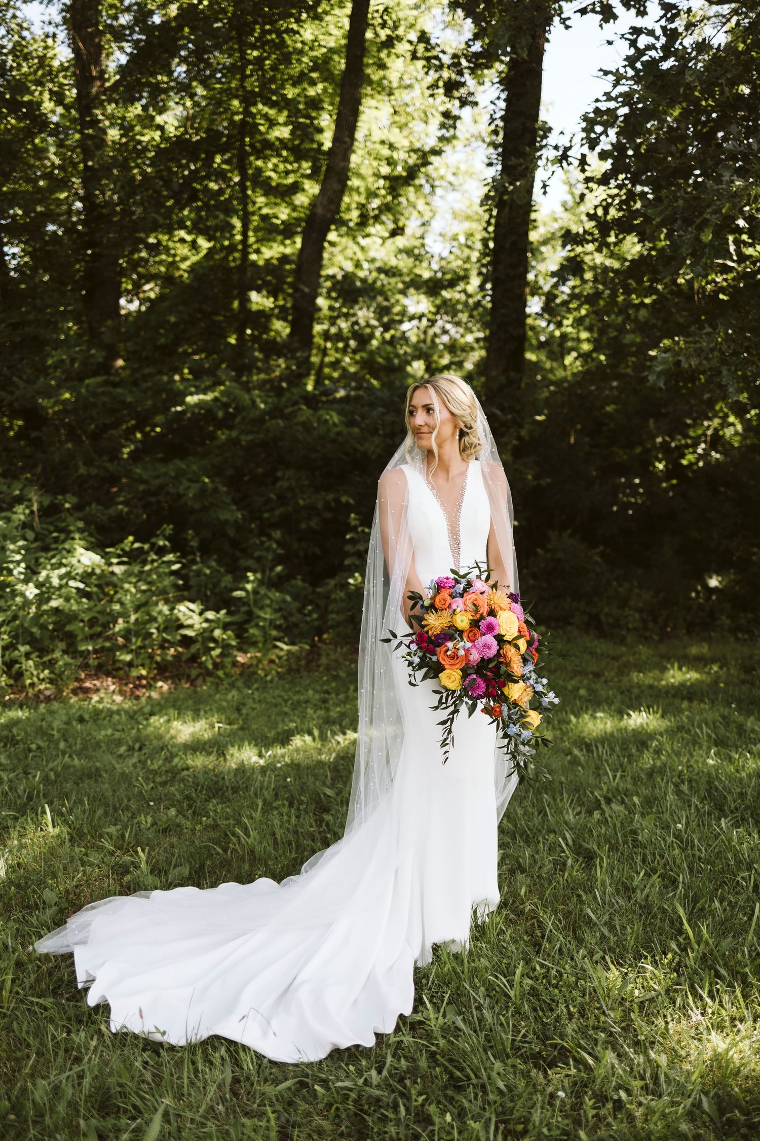 Elegant bride stands in sunlit forest wearing plunging neckline gown and holding colorful cascading bouquet.