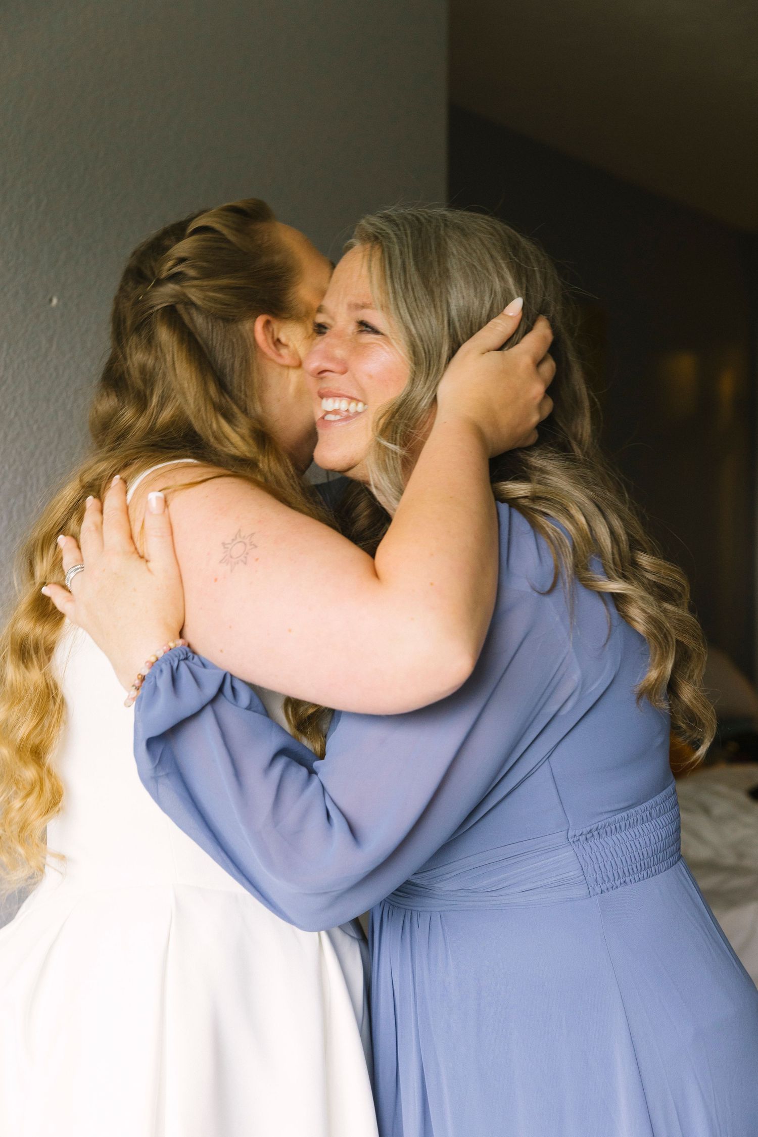 Two women embrace in an emotional moment, one wearing a white dress and the other in a flowing periwinkle blue gown.