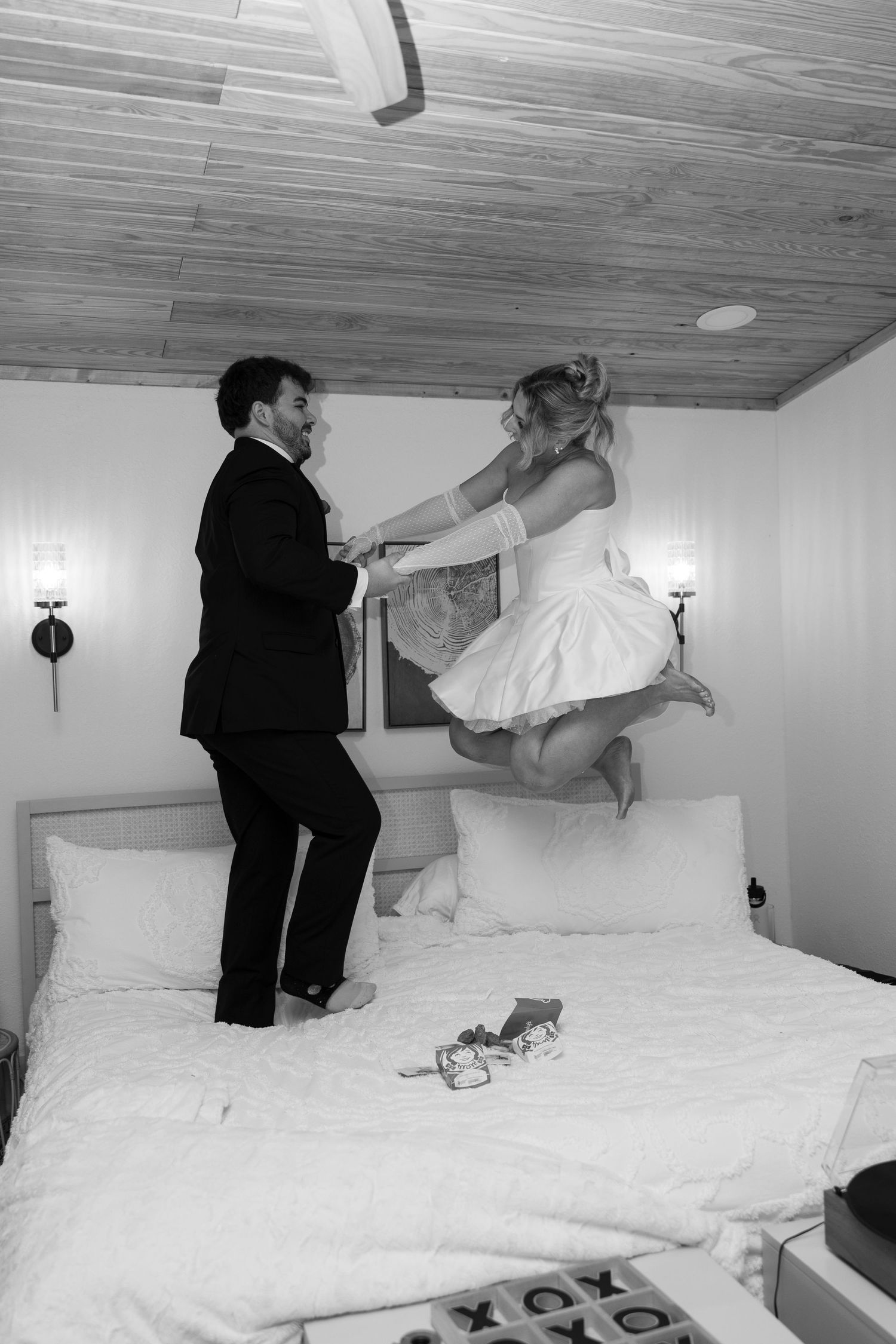 A black and white photo of a couple in wedding attire playfully jumping on a bed with white linens.