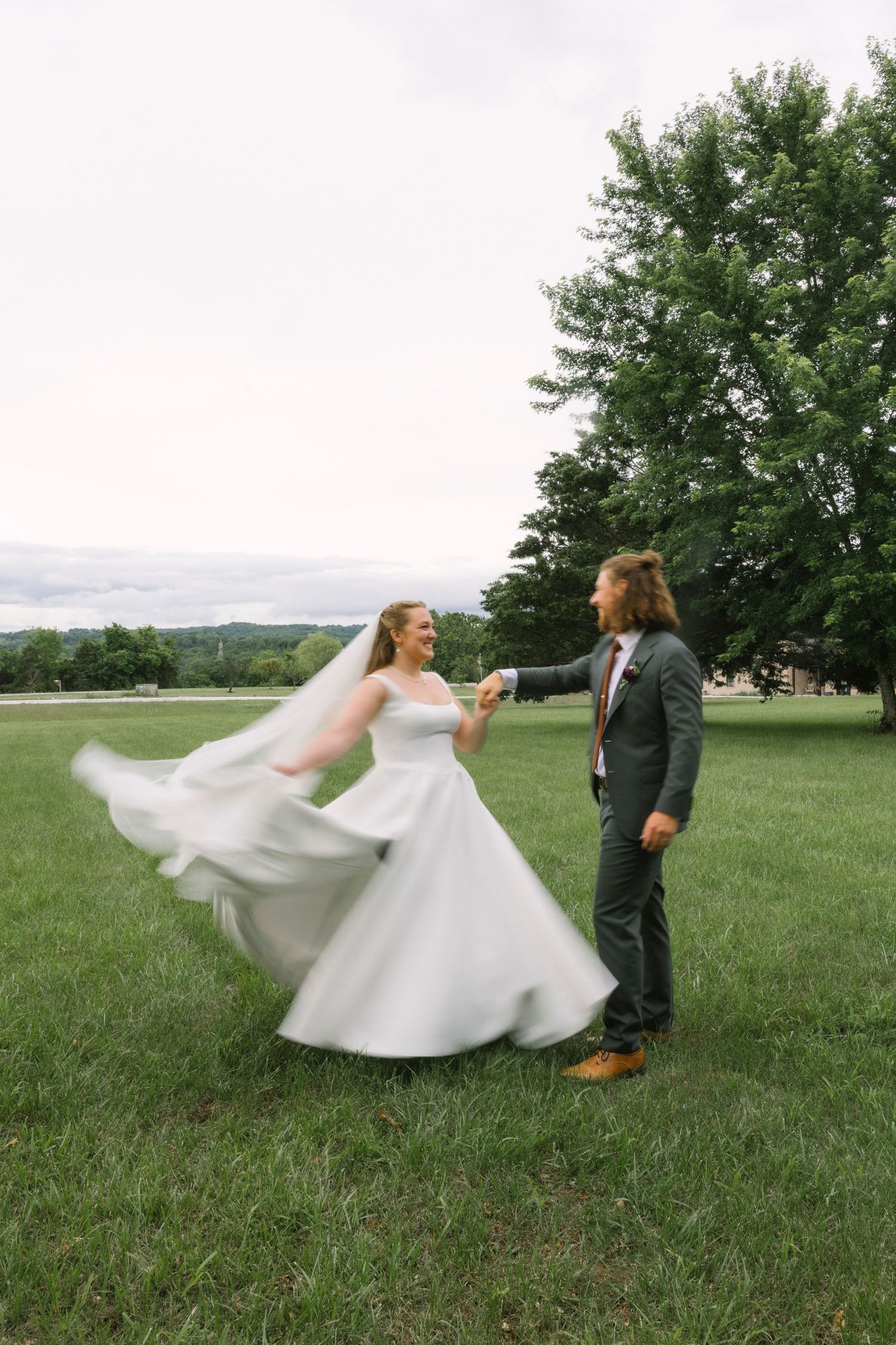 A bride twirls her dress while sharing a joyful moment with her partner in a scenic meadow.
